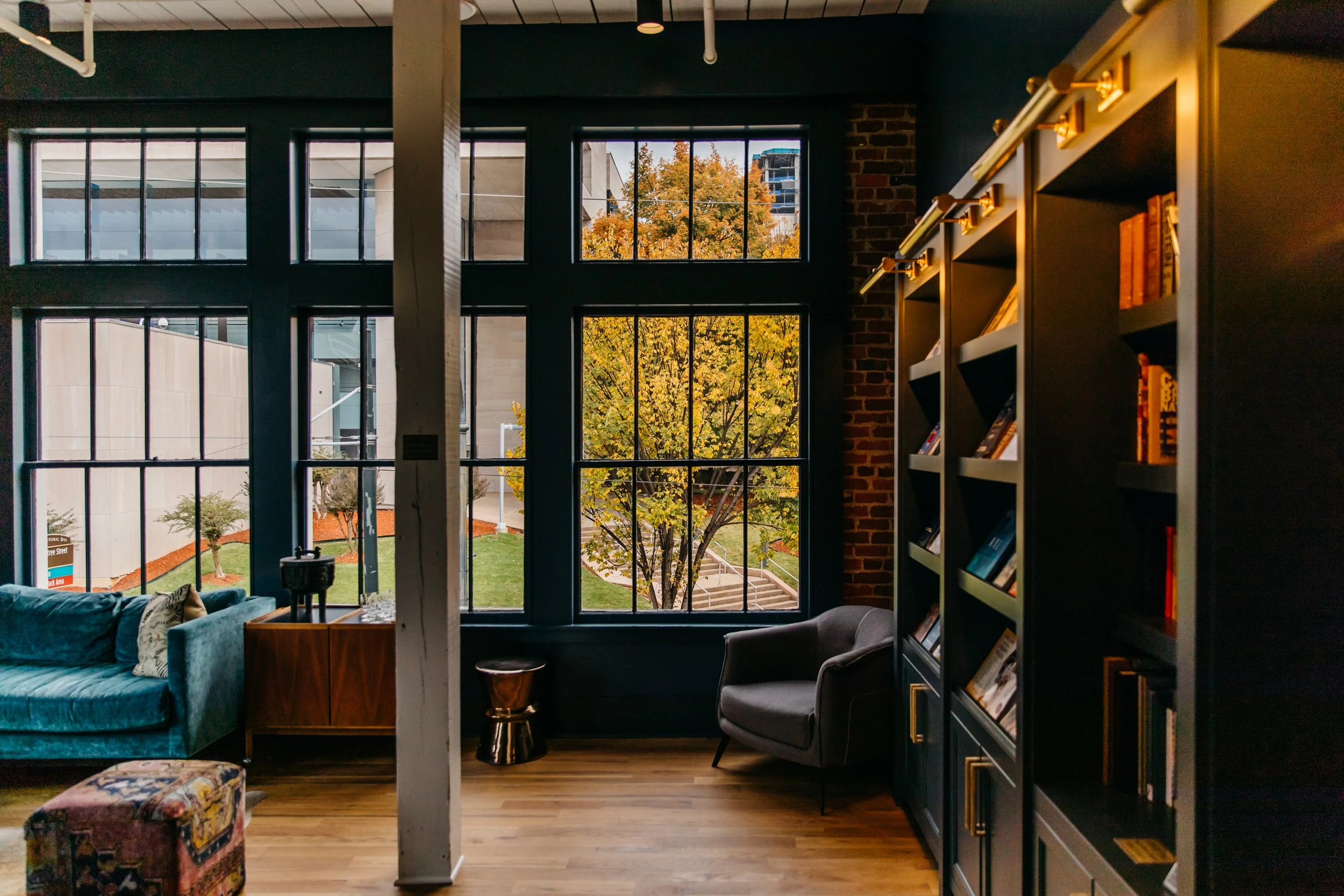 Interior of a modern room with large grid-patterned windows showing trees with autumn foliage outside. On the left, there is a blue velvet sofa with pillows, and on the right, a bookshelf with books. A small table and a chair are also visible, with a