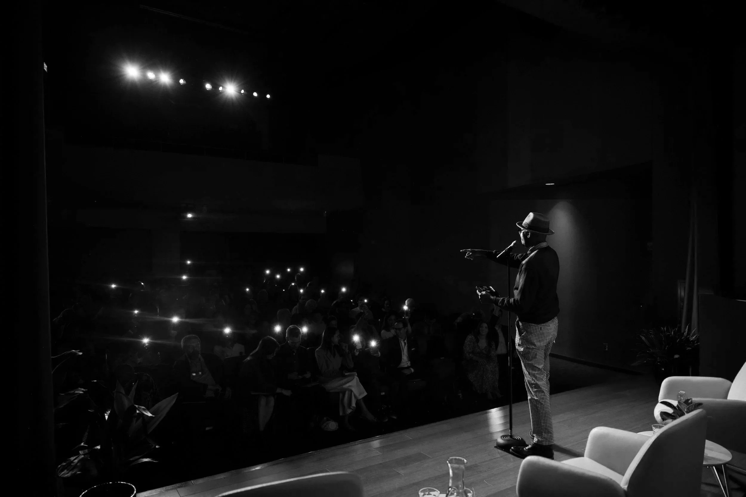 A man in a hat and checkered pants stands on stage speaking into a microphone in a dark auditorium. The audience is visible with some people using their phones' flashlight features. The stage is minimally lit, and the background is mostly dark.