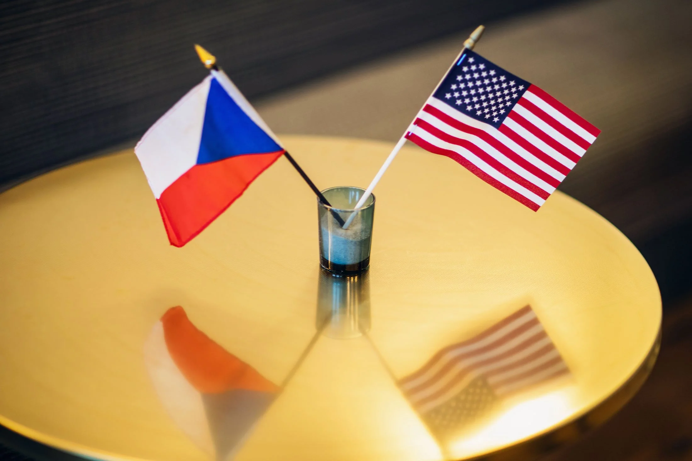 Two small flags, one of the United States and one of the Czech Republic, are placed in a clear glass on a yellow table. The flags are positioned close to each other.