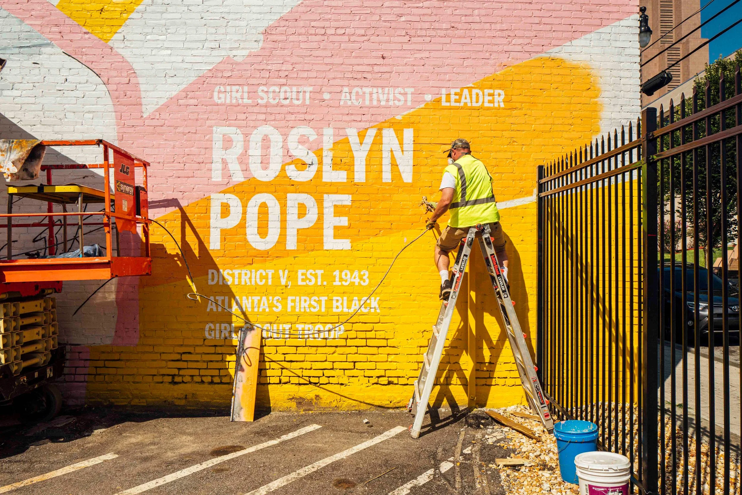 A person wearing a yellow safety vest and cap painting a mural on a brick wall with yellow and pink sections. The mural features white text reading 'Girl Scout · Activist · Leader · Roslyn Pope · District V, Est. 1943 · Atlanta's First Black Girl Scout Troop.' Tools and buckets are on the ground, and a lift is present on the left side.