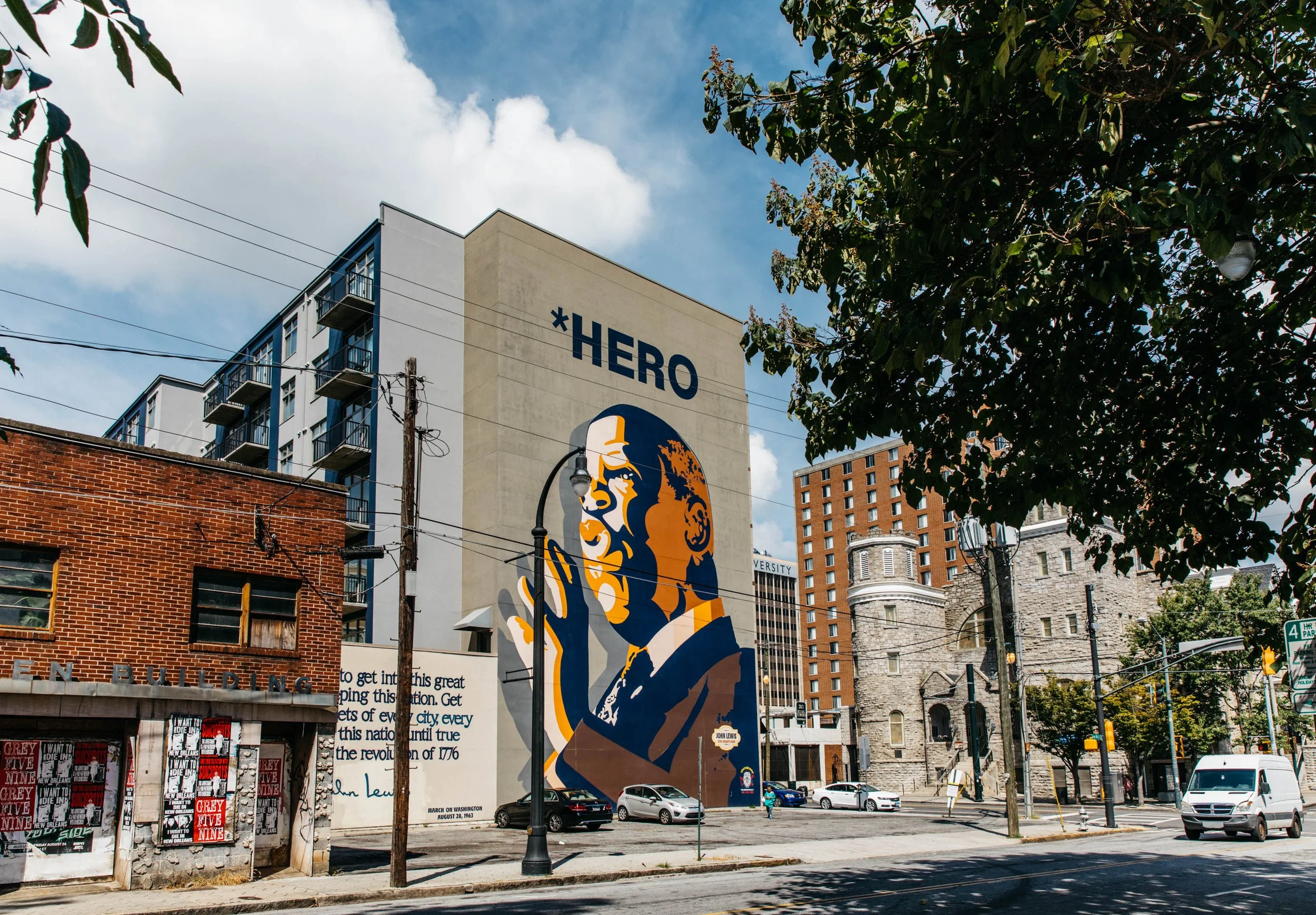 A city street scene with a large mural of a historical figure, the word "HERO" on a building, and various buildings, trees, and cars parked along the street.