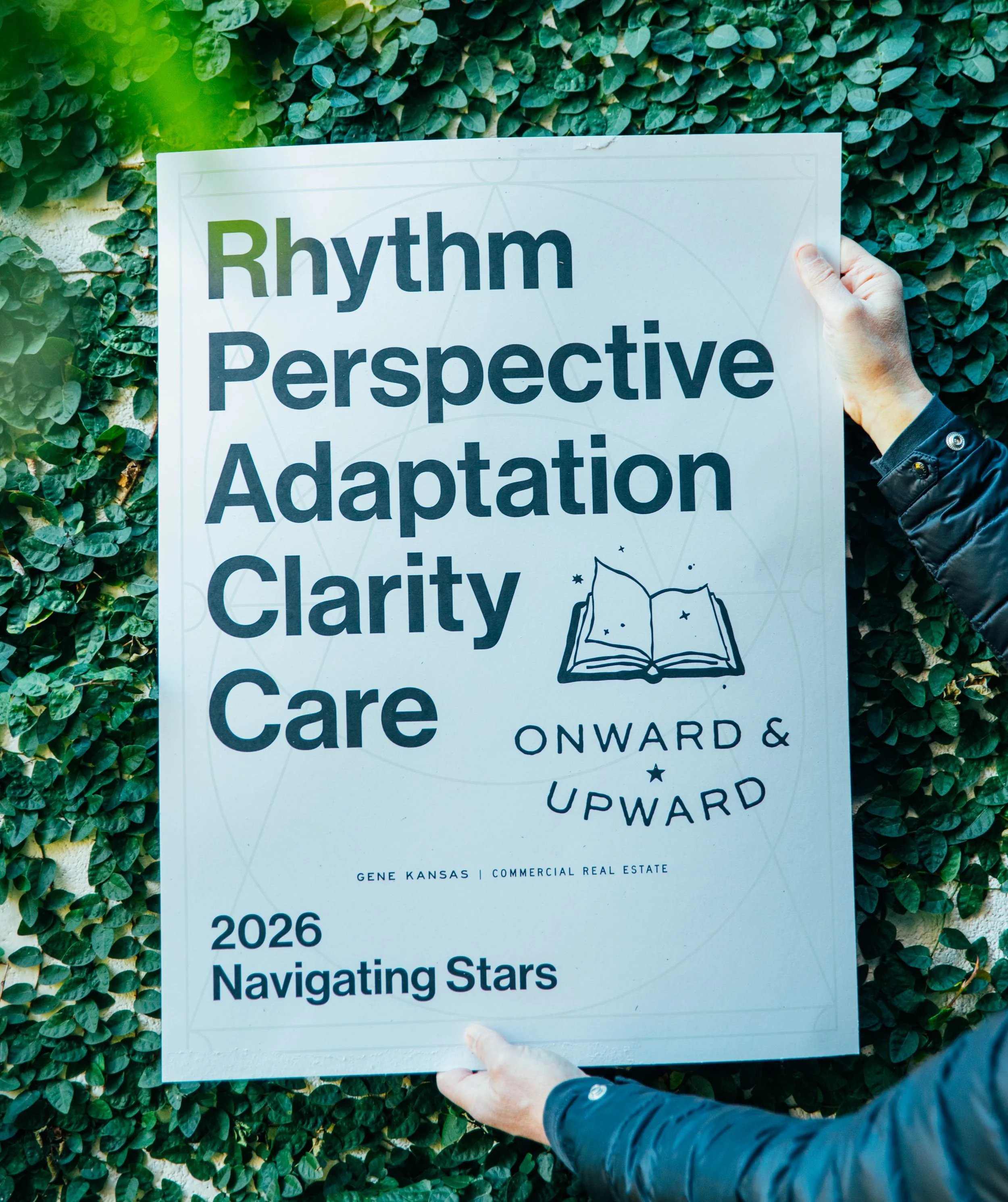 Person holding a white sign with black and green text, surrounded by green foliage. The sign features the words "Rhythm Perspective Adaptation Clarity Care" with a small illustration of an open book.