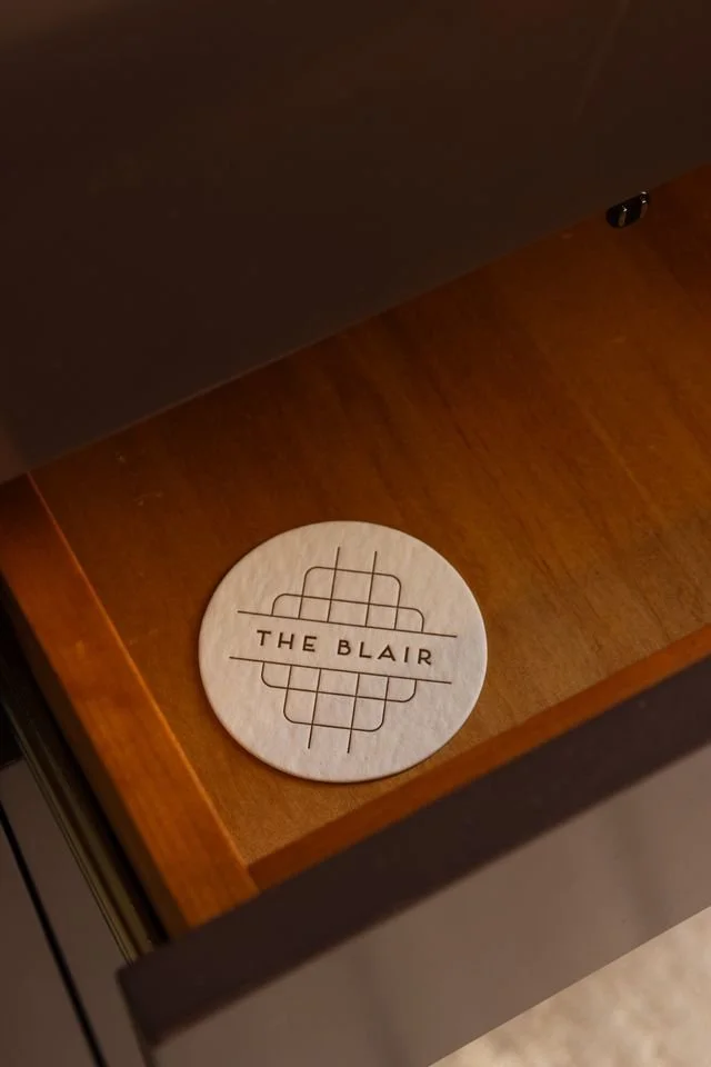 Circular wooden coaster with the logo and text 'THE BLAIR' on a wooden table.