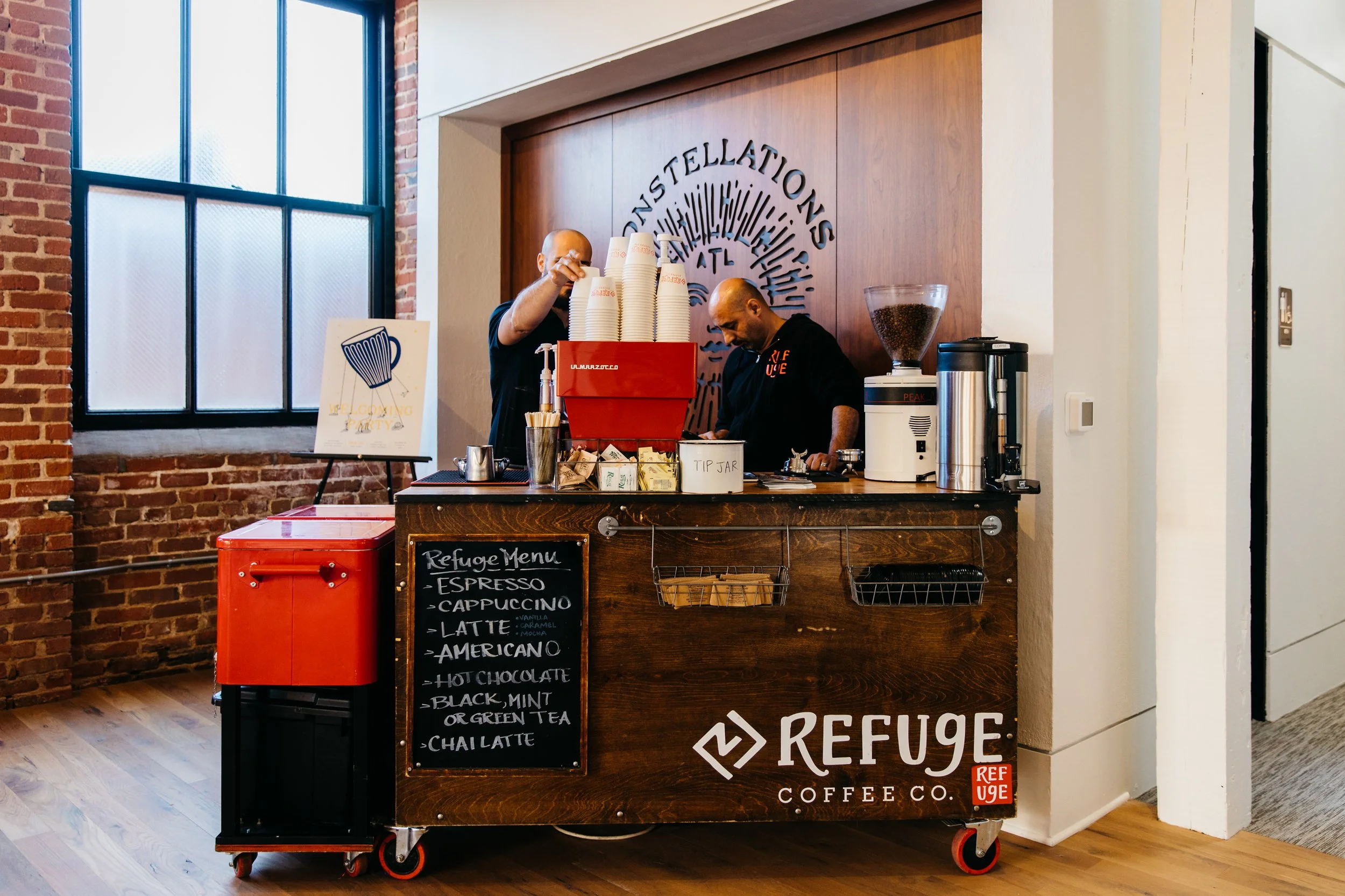 A coffee cart with a blackboard menu listing various coffee drinks, a tip jar, and cups stacked on top of a red espresso machine. Behind the cart, two baristas are preparing coffee. The setting is an indoor café with brick walls, a large window, and wooden floors.