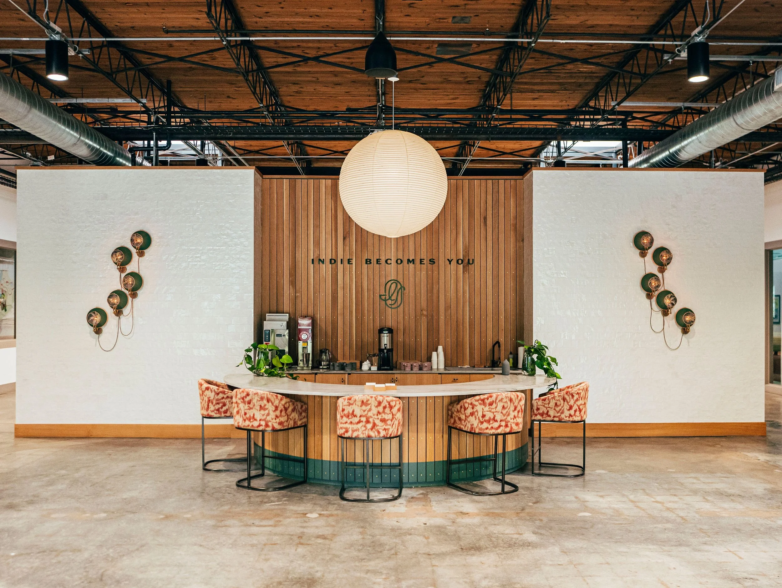 A modern coffee shop counter with a large paper lantern hanging above, surrounded by orange and pink patterned chairs. The background features a wood-paneled wall with the phrase 'Indie Becomes You' and a minimalist logo.