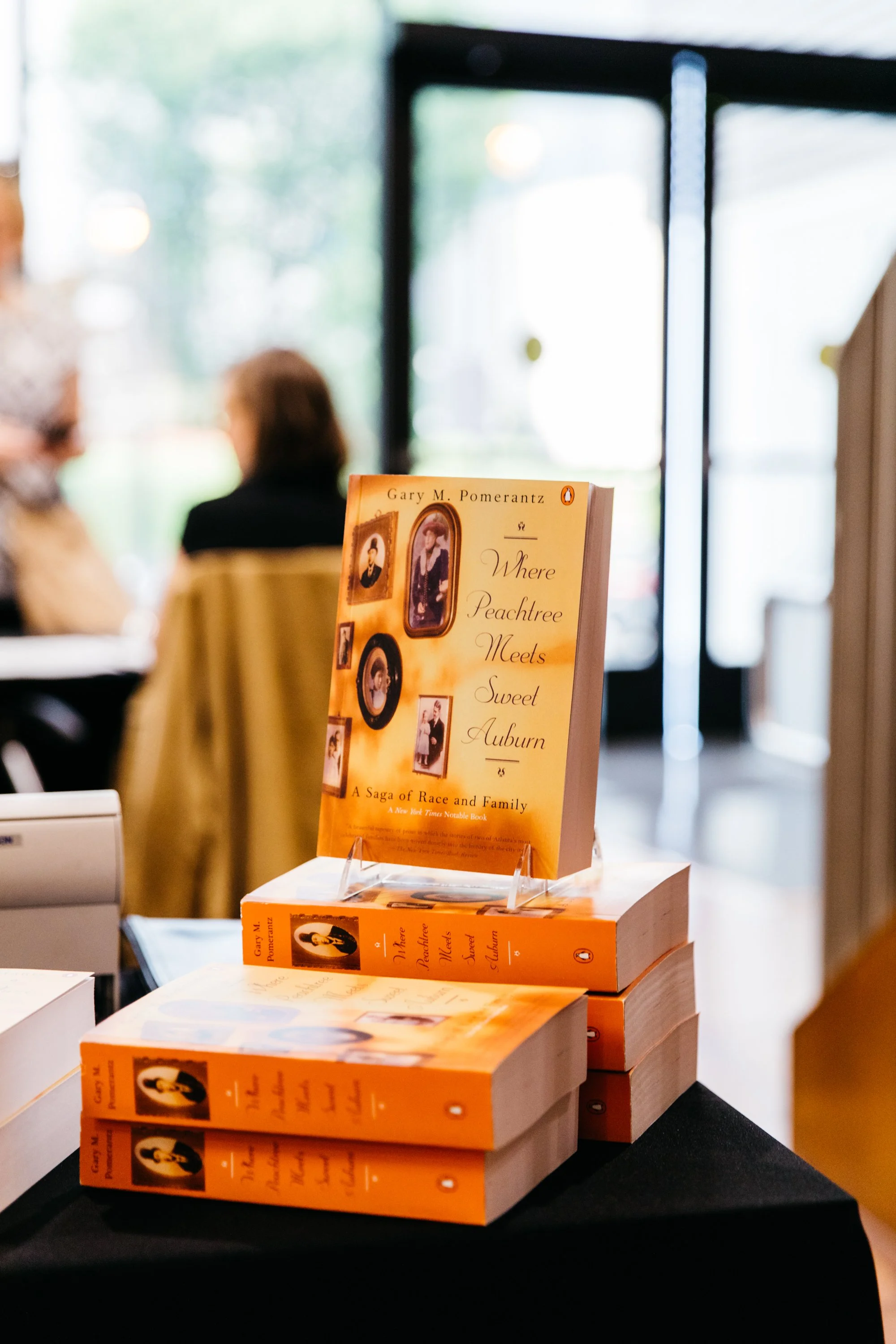 Stack of orange books titled "Where Peachtree Meets Sweet Auburn" by Gary M. Pomerantz, displayed on a table inside a bookstore or cafe.