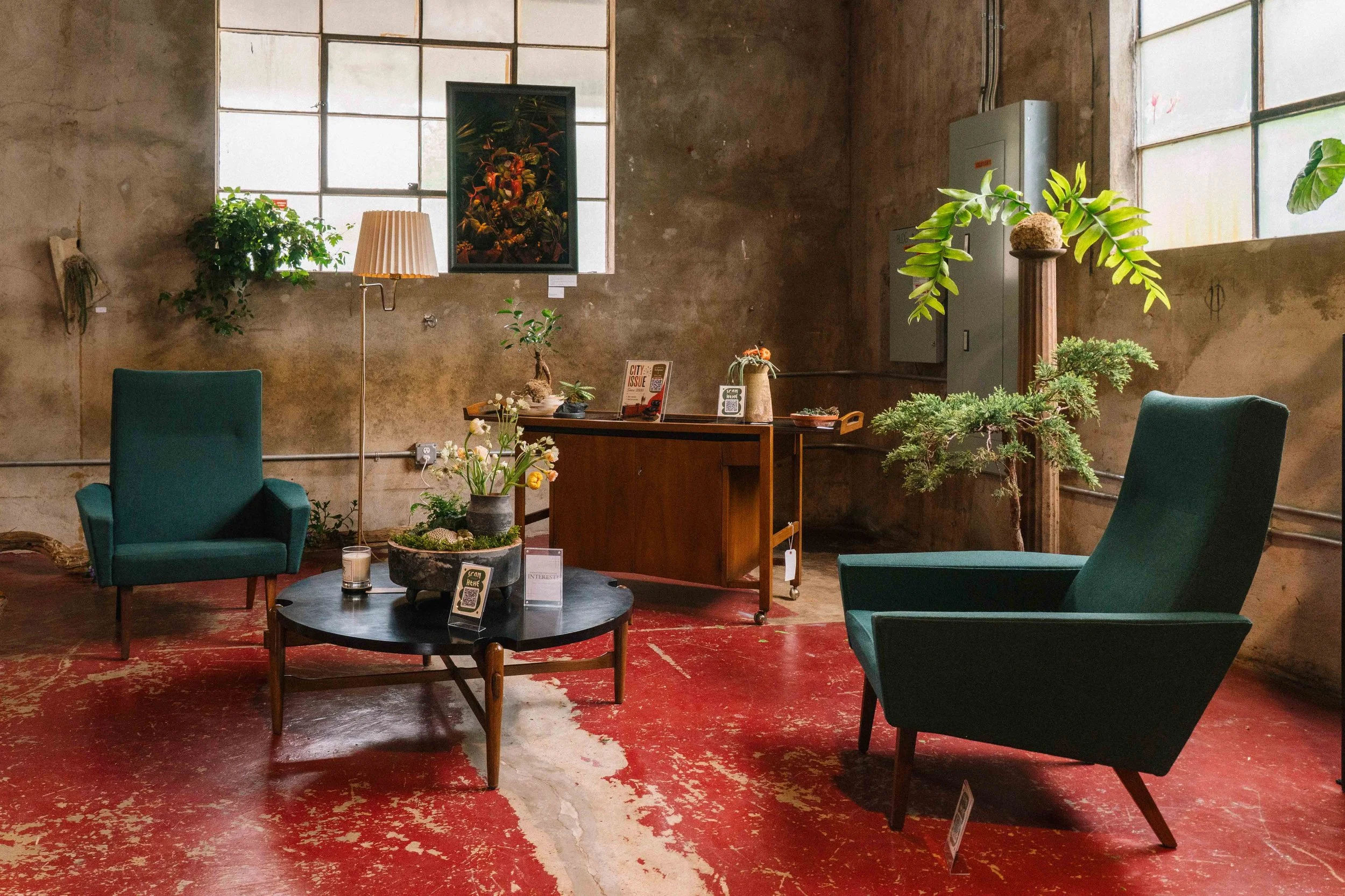 Indoor space with a red and worn wooden floor, two teal armchairs, a round black coffee table with small potted plants, decorative items, and informational signs, a wooden cabinet with plants and magazines, green plants on the wall and in pots, a fra
