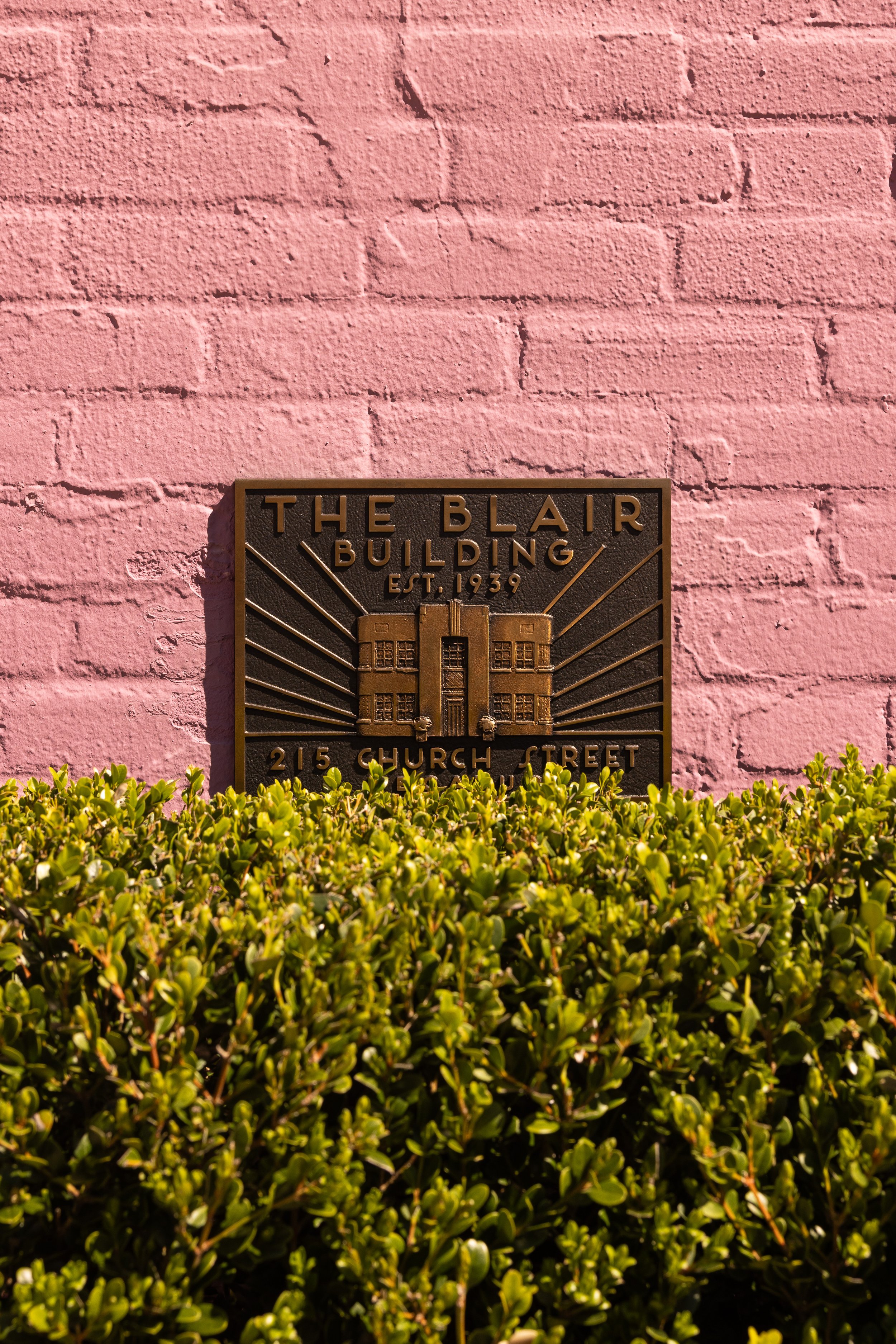 Plaque on a pink brick wall reads 'The Blair Building Est. 1939, 215 Church Street' with green bushes in the foreground.