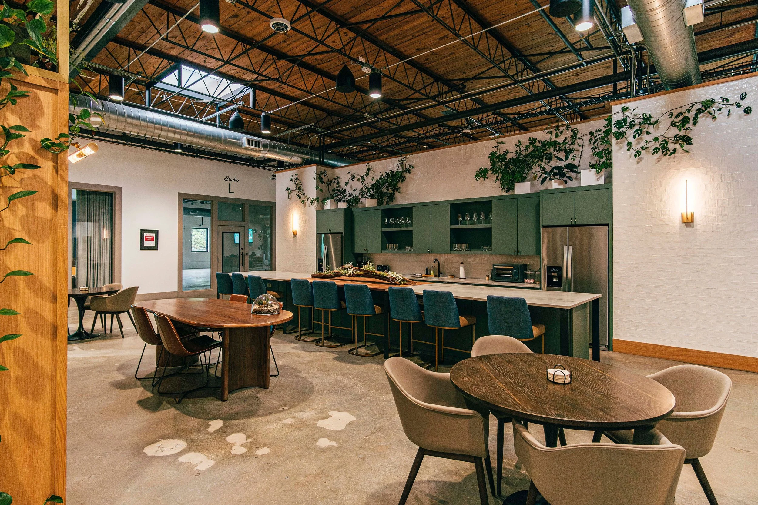 Modern indoor restaurant or cafe with wooden ceiling, green cabinets, plants, and tables with chairs.