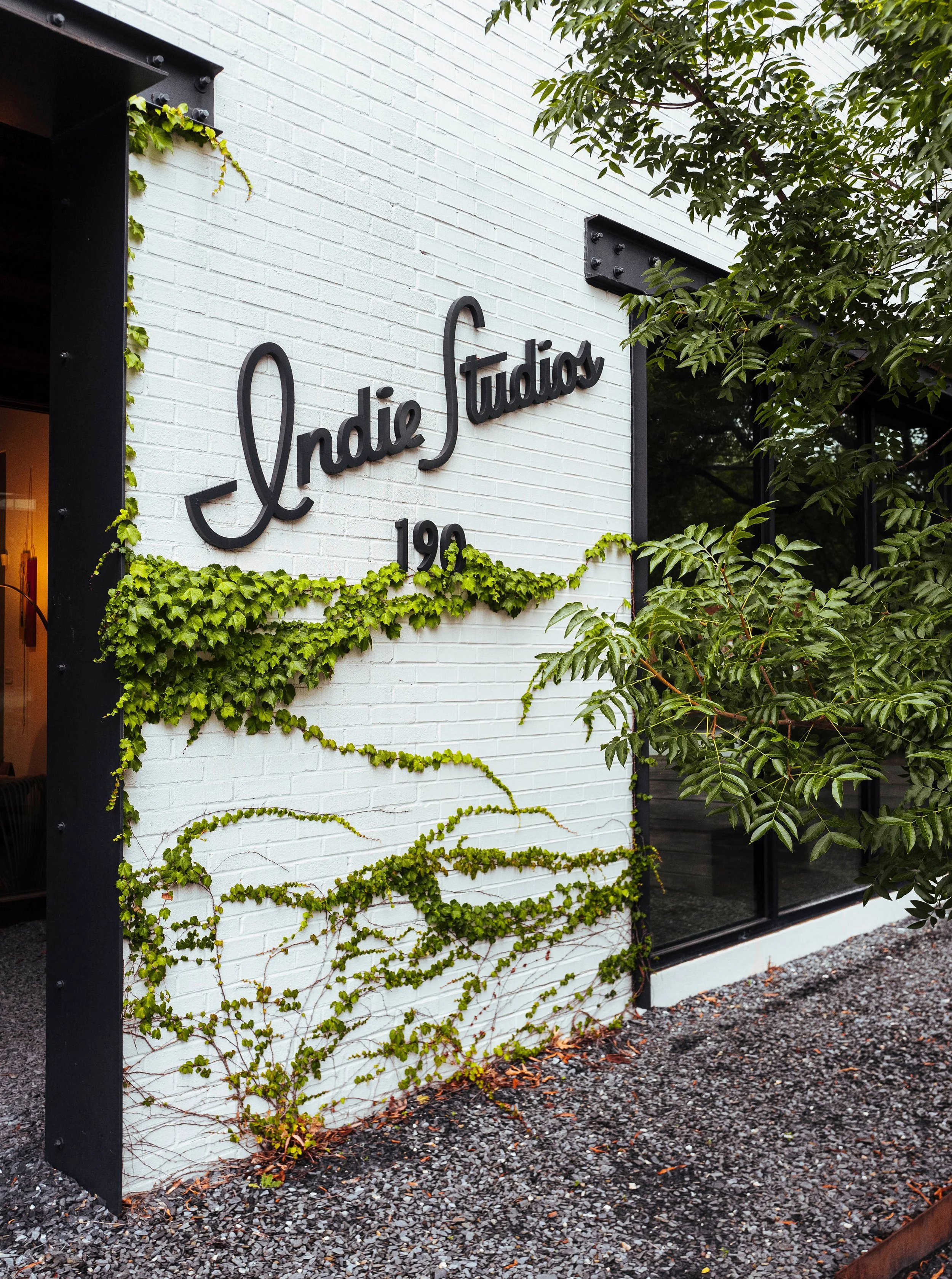 A white brick wall with black metal signage that reads 'Indie Studios 190', surrounded by green ivy and nearby trees, and a gravel path at the bottom.