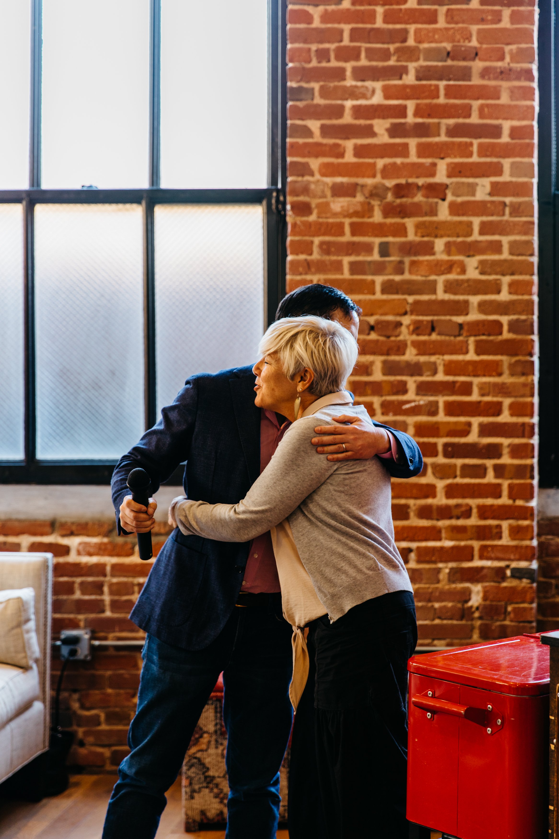 Two people hugging in an industrial-style room with brick walls and large windows, one holding a microphone.