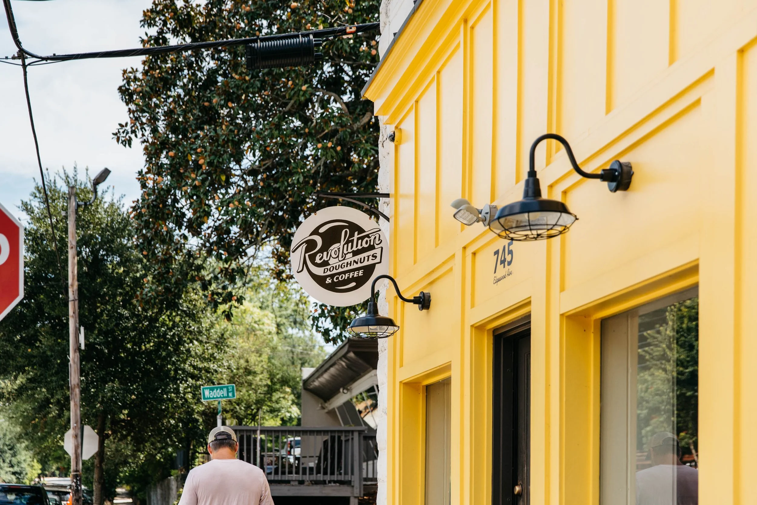 Yellow building with black light fixtures and a sign for Revolution Doughnuts & Coffee, with trees and a man in a backwards cap walking in front.
