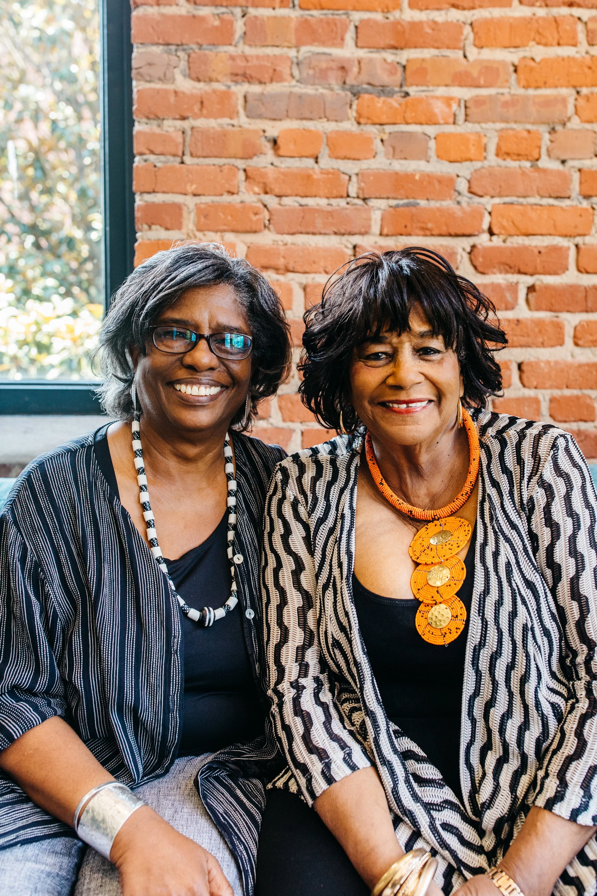 Two women sitting closely together in front of a brick wall, smiling at the camera.