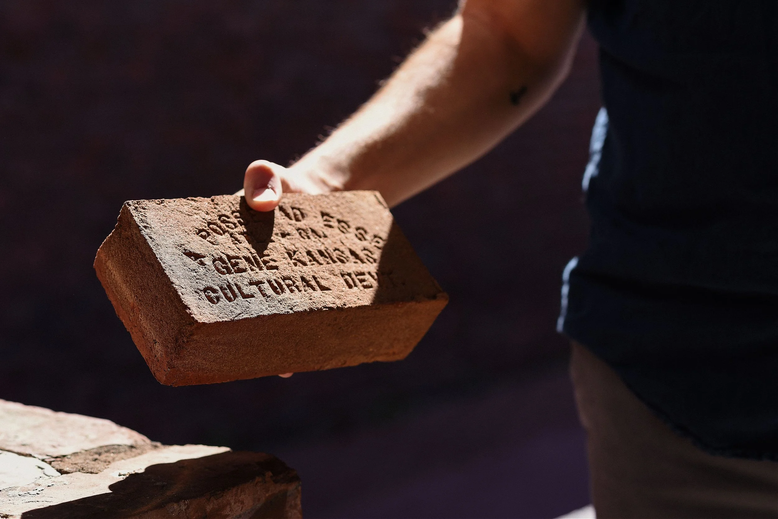 Person holding a brick with embossed text, part of a structure or construction site, dark background.