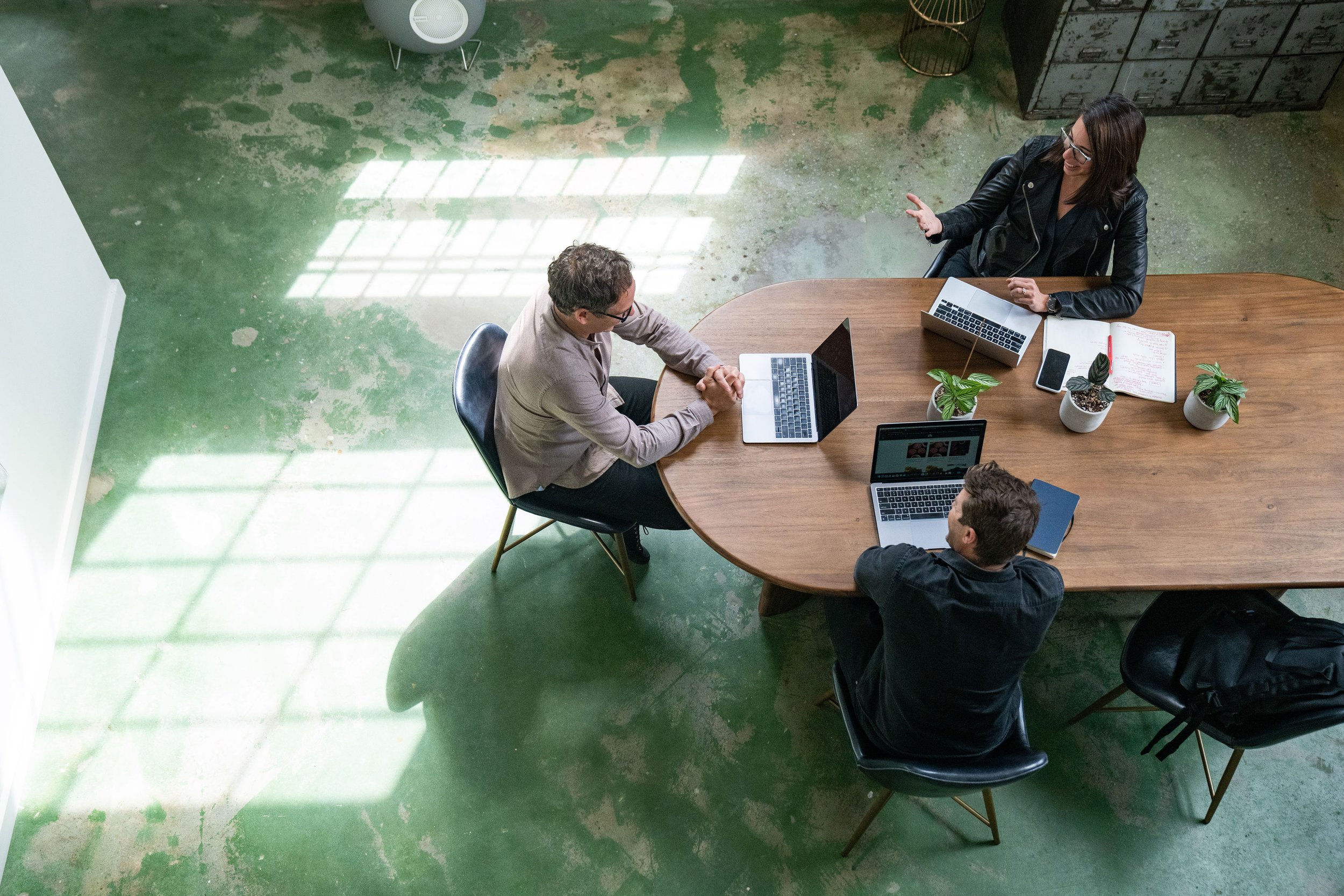 Overhead view of three people sitting at a wooden conference table having a discussion in a room with a green concrete floor and large windows creating bright reflections on the floor.