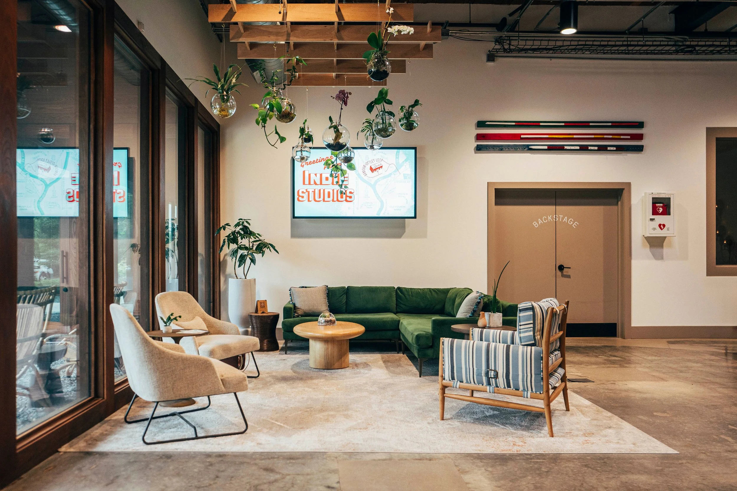 A cozy lounge area with a green sofa, single armchairs, and a small wooden coffee table, decorated with indoor plants and a large wall-mounted monitor displaying a logo for 'Indie Studios'. Hanging plants are suspended from a wooden ceiling structure