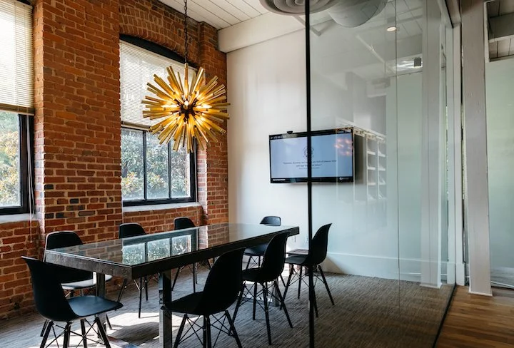 Modern conference room with brick walls, large windows, a glass-top table, six black chairs, a gold starburst chandelier, and a wall-mounted flat-screen TV.