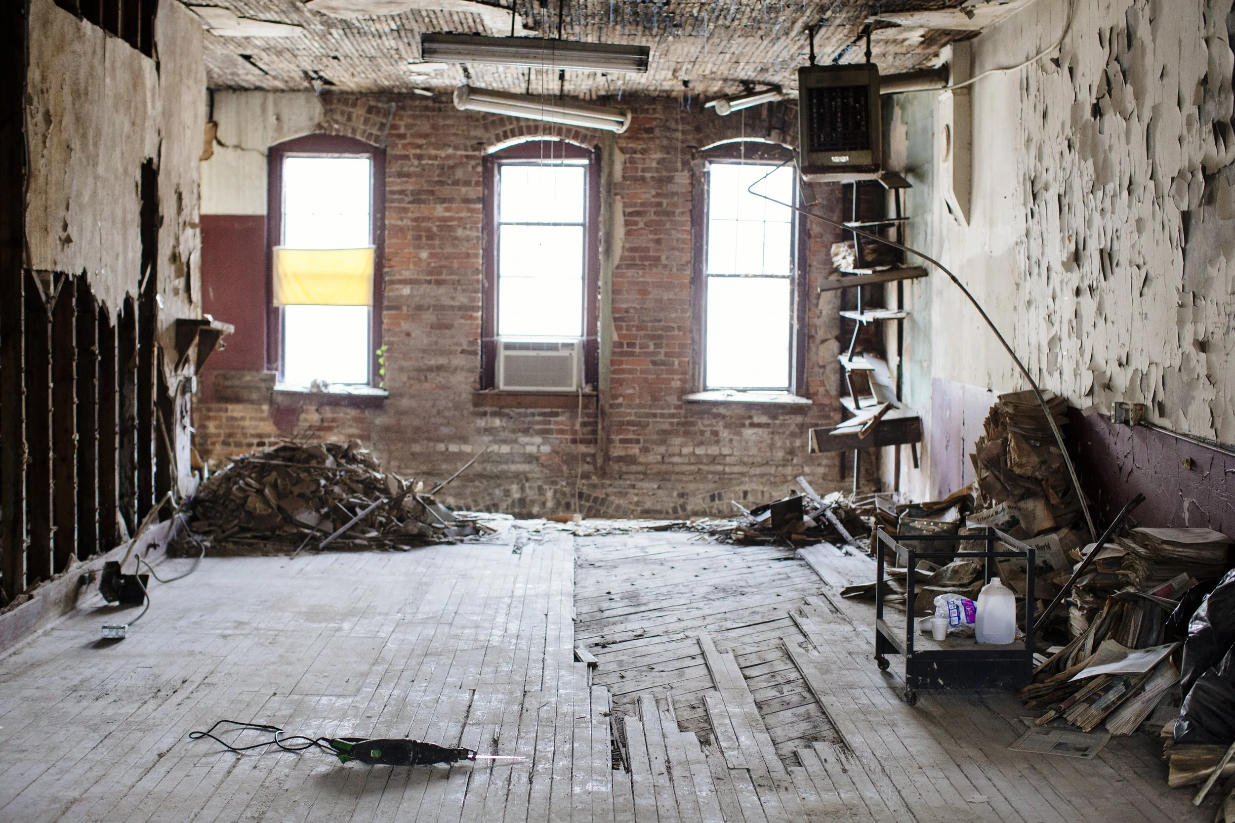 An abandoned, partially demolished room with three large windows, exposed brick walls, and debris on the floor, including wood, paper, and trash.