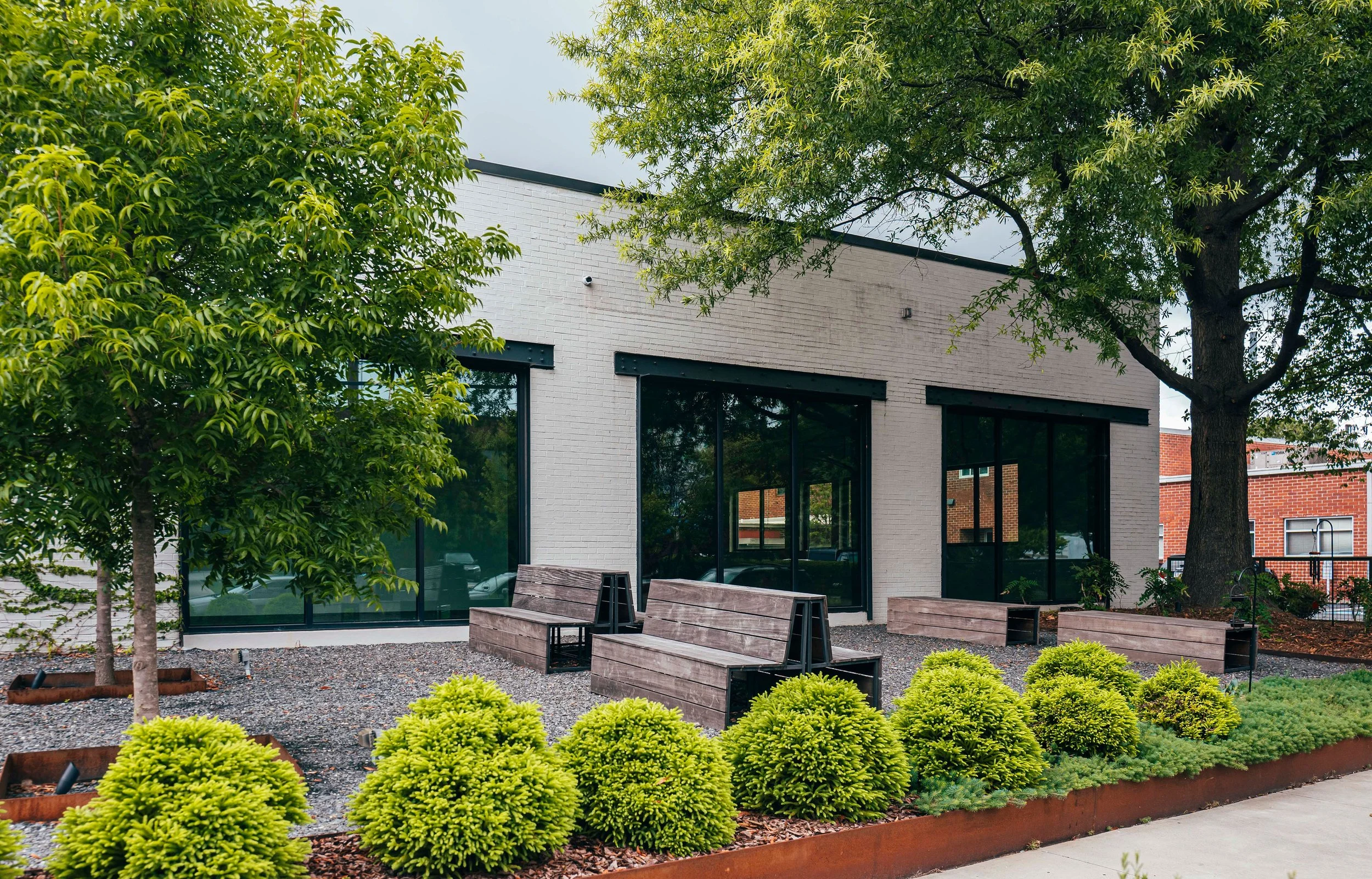Modern building with large glass windows, surrounded by trees and manicured shrubs, with outdoor benches and gravel on the ground.