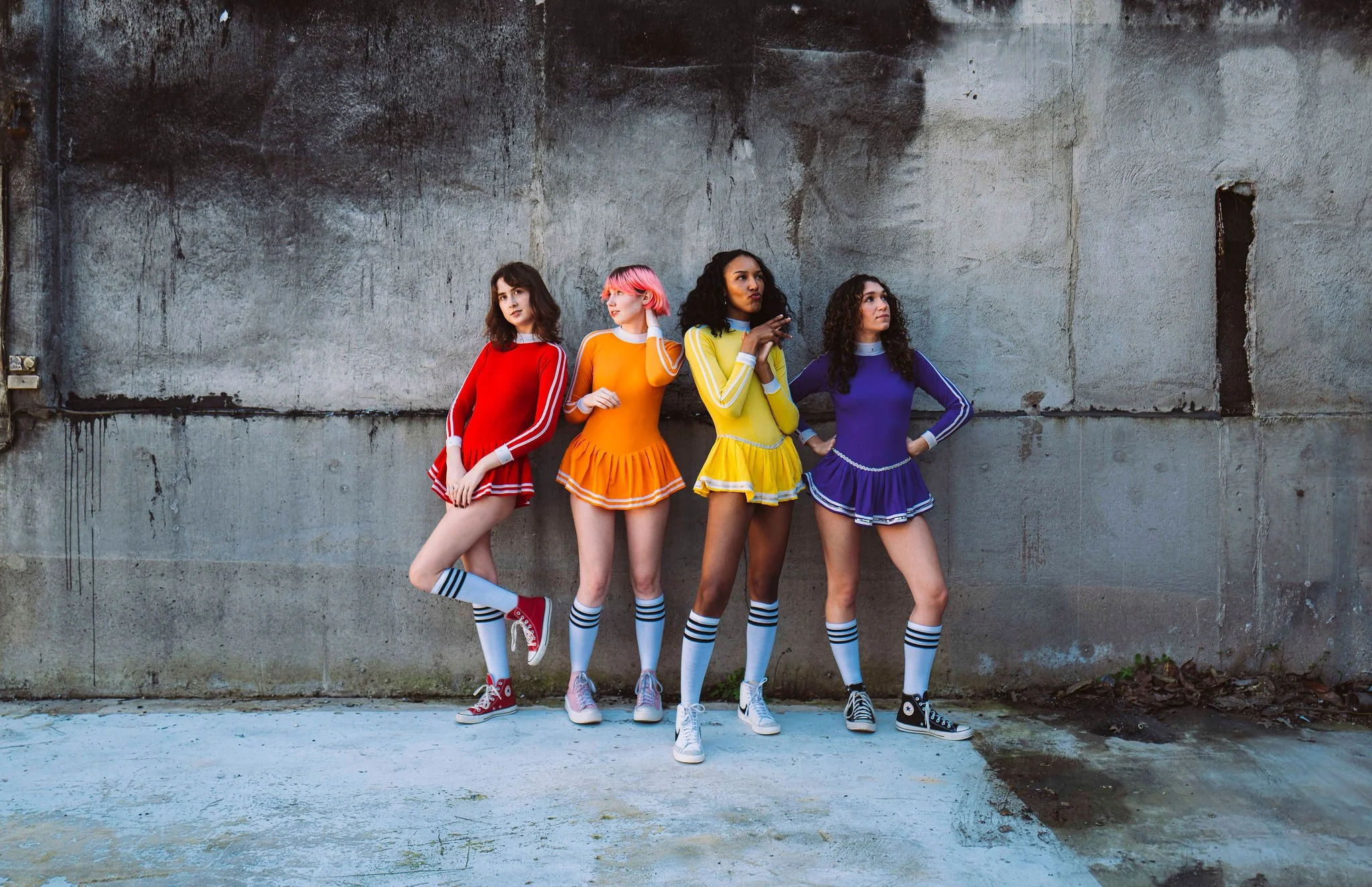 Four women stand against a concrete wall, wearing colorful cheerleading outfits in red, orange, yellow, and purple, with matching striped knee-high socks and sneakers.