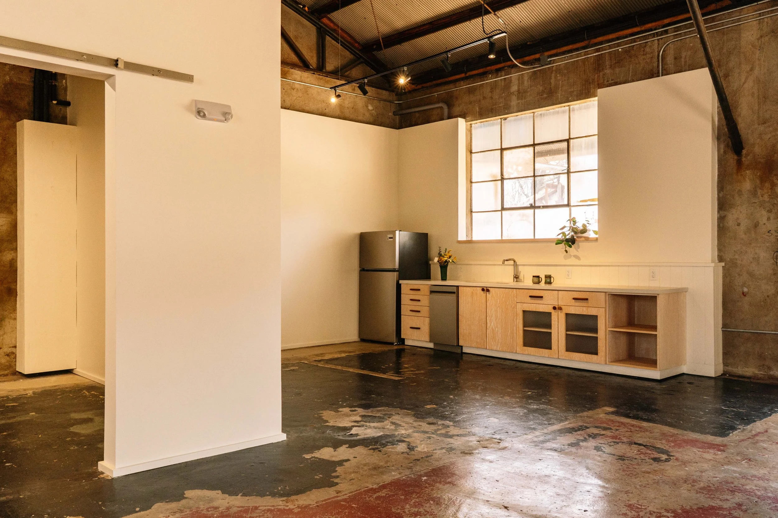 Interior of an industrial-style kitchen with exposed brick walls, large window, wooden cabinets, and a black refrigerator. The ceiling has visible pipes and track lighting, and the floor is worn with patches of paint.
