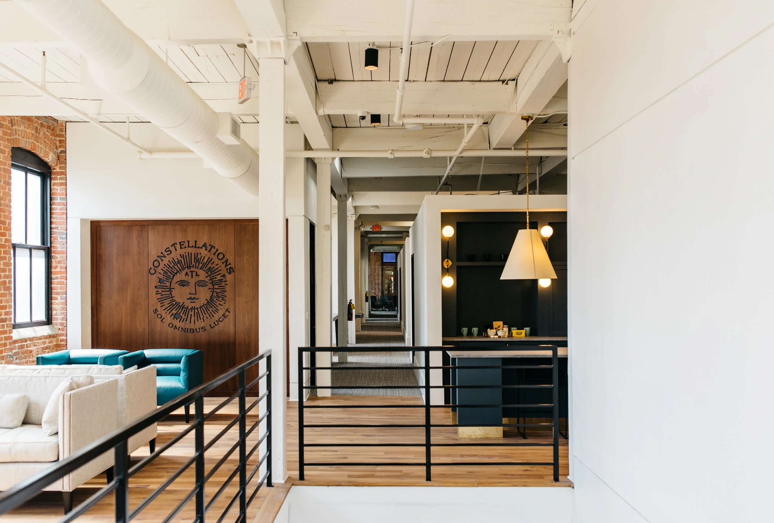 Interior of a modern apartment or office with white walls and exposed brick walls, featuring black and white furniture, hanging lights, windows, and a staircase with a black railing.