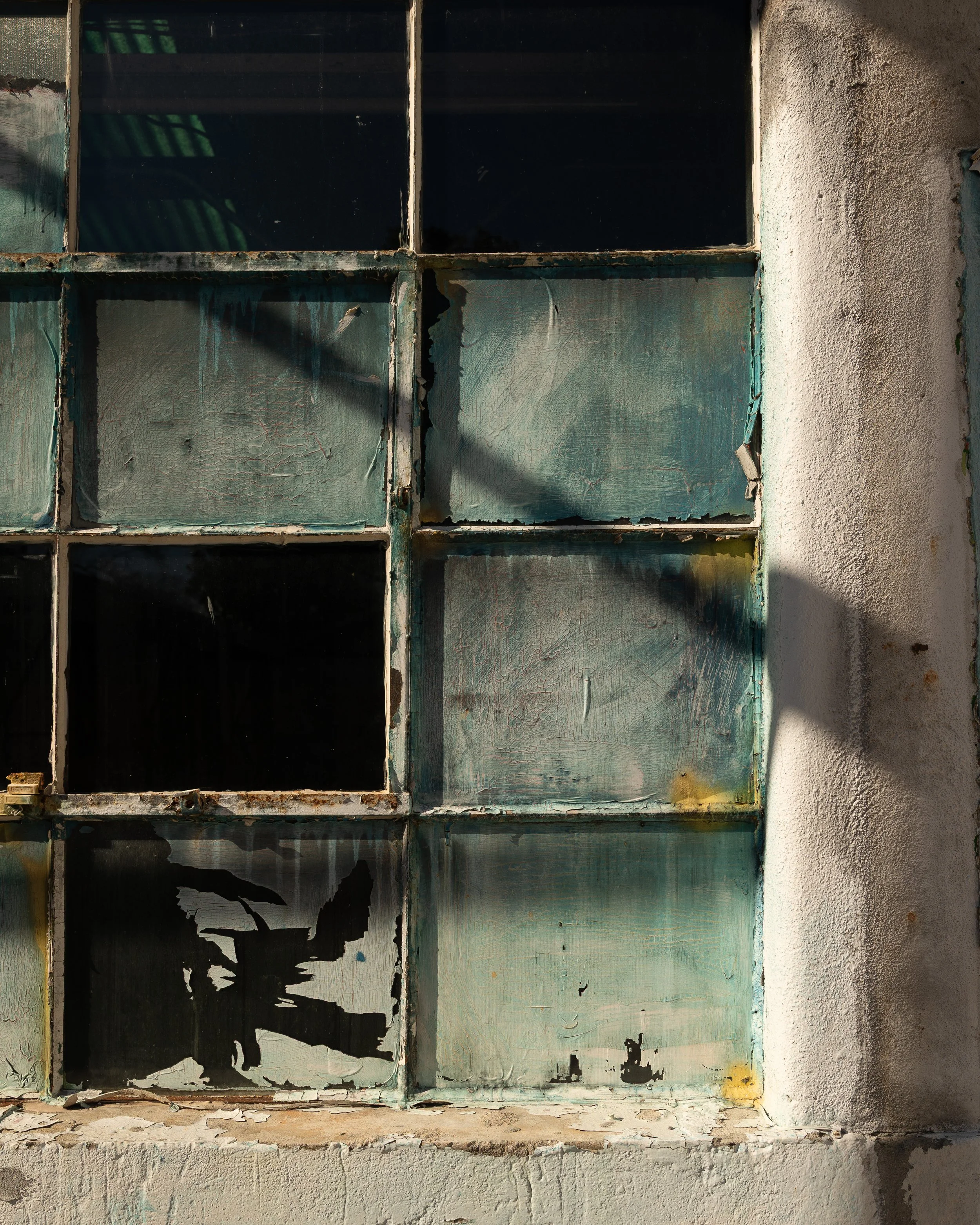 A window with old, painted glass panes and rusted metal frames set in a weathered wall with peeling paint and cracks.