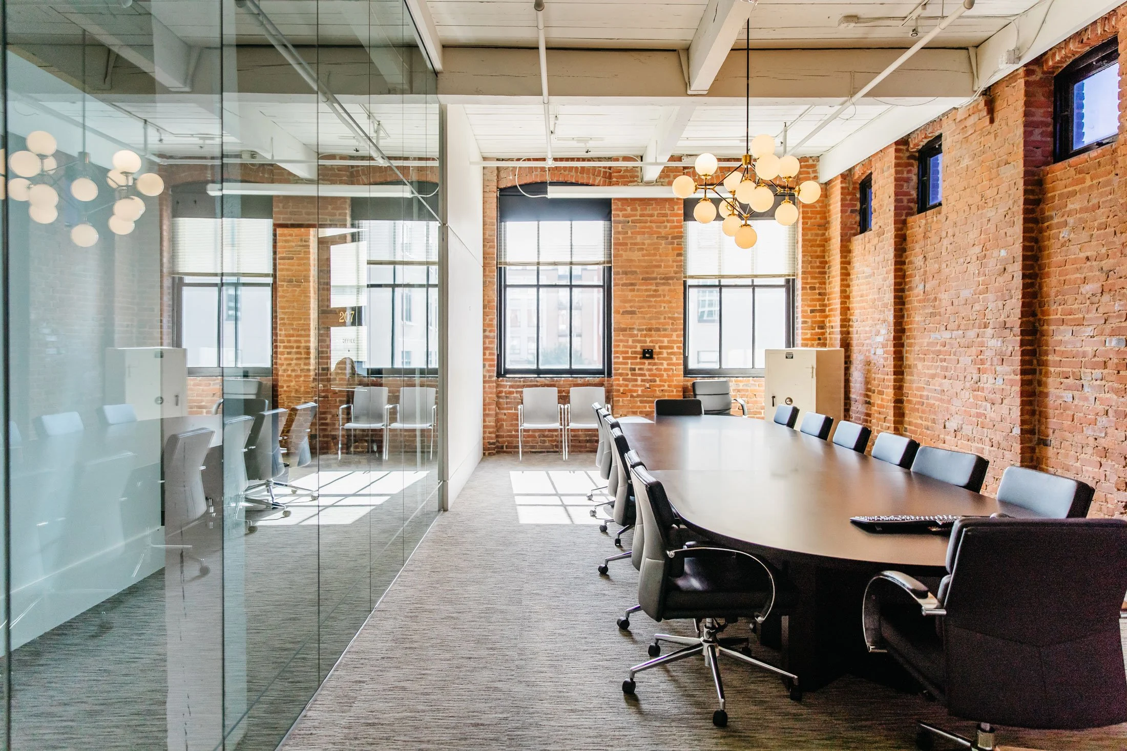 A modern conference room with exposed brick walls, large windows with blinds, and a long conference table surrounded by black office chairs. There are ceiling-mounted light fixtures and a glass wall dividing the room. Sunlight streams through the win