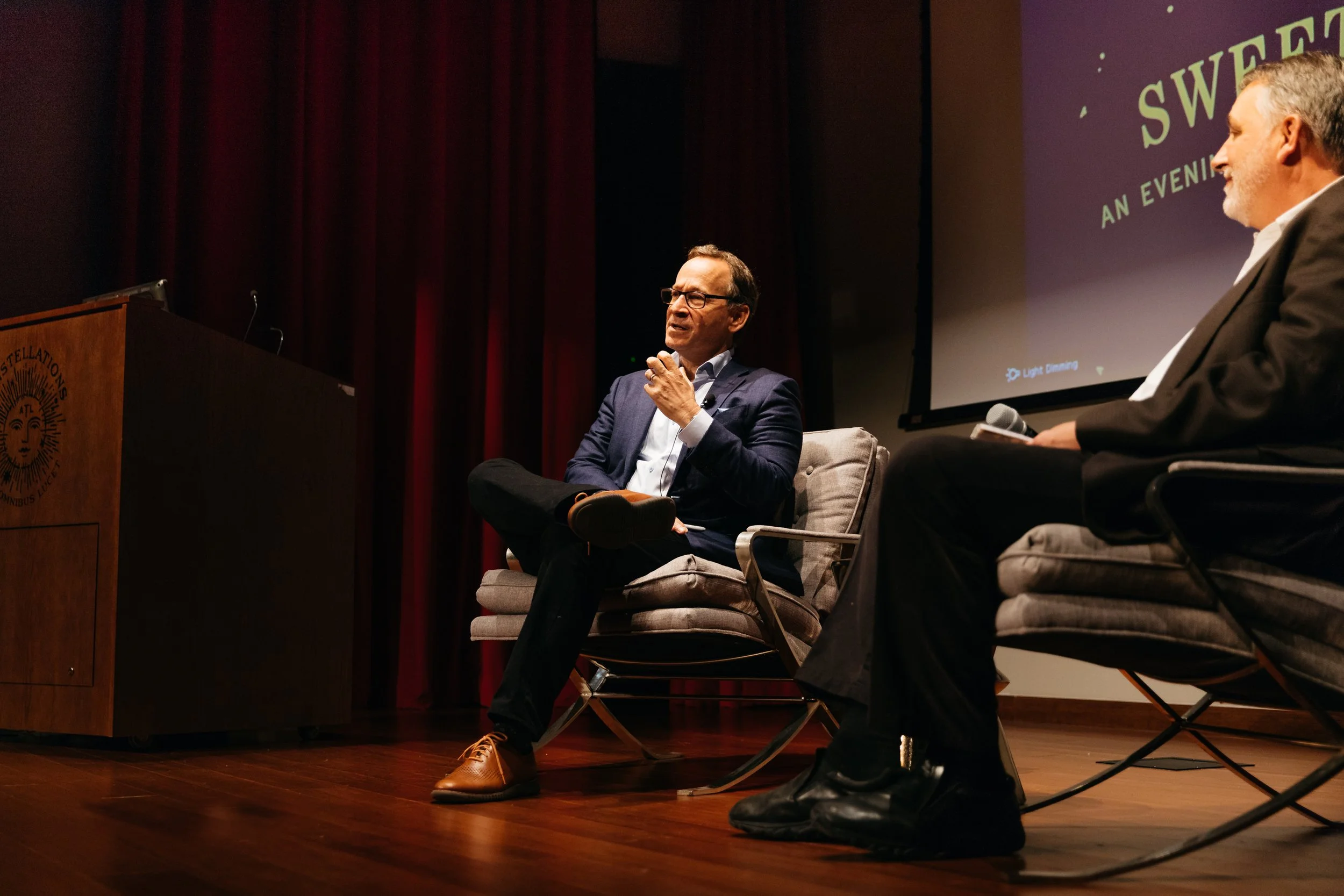 Two men are seated on stage during a panel discussion at an event. One man is speaking while the other listens. A large screen behind them displays a presentation. The setting appears formal, with a wooden podium on the left and dark curtains in the background.