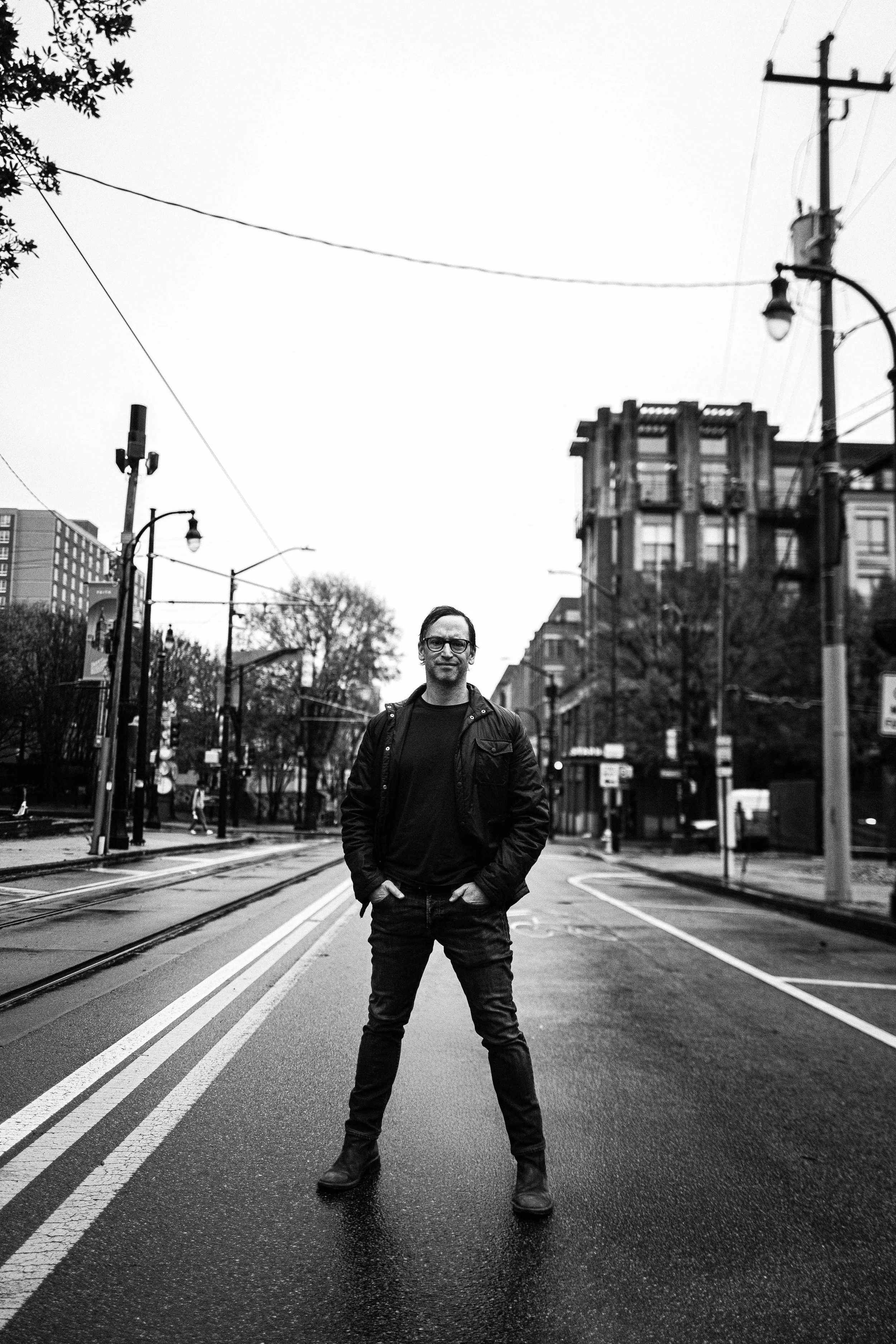 A man standing in the middle of a city street with tram tracks, wearing glasses, a black jacket, and jeans, with buildings, street lamps, and power lines in the background.