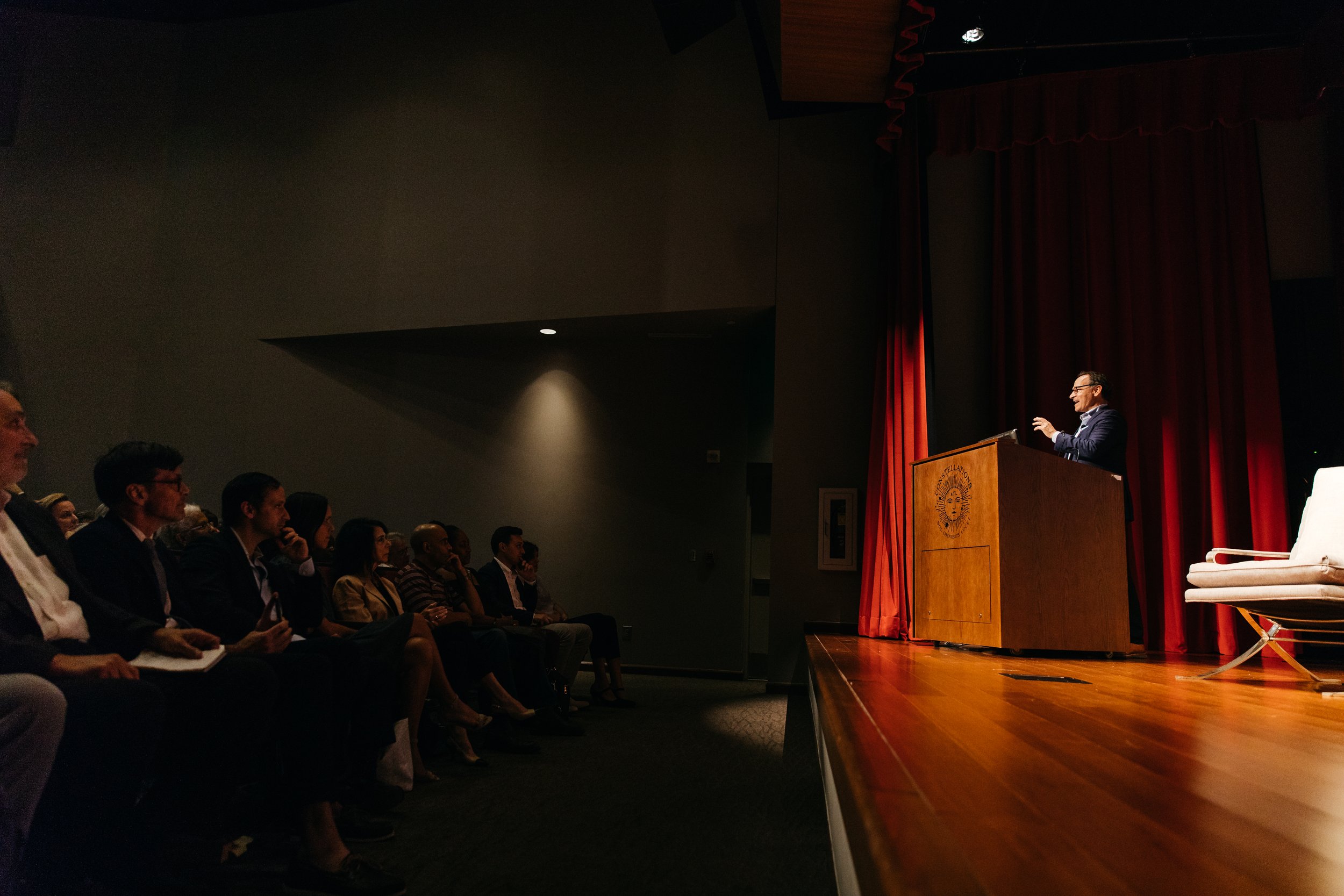 Man in a suit giving a speech at a podium in an auditorium with an audience watching.