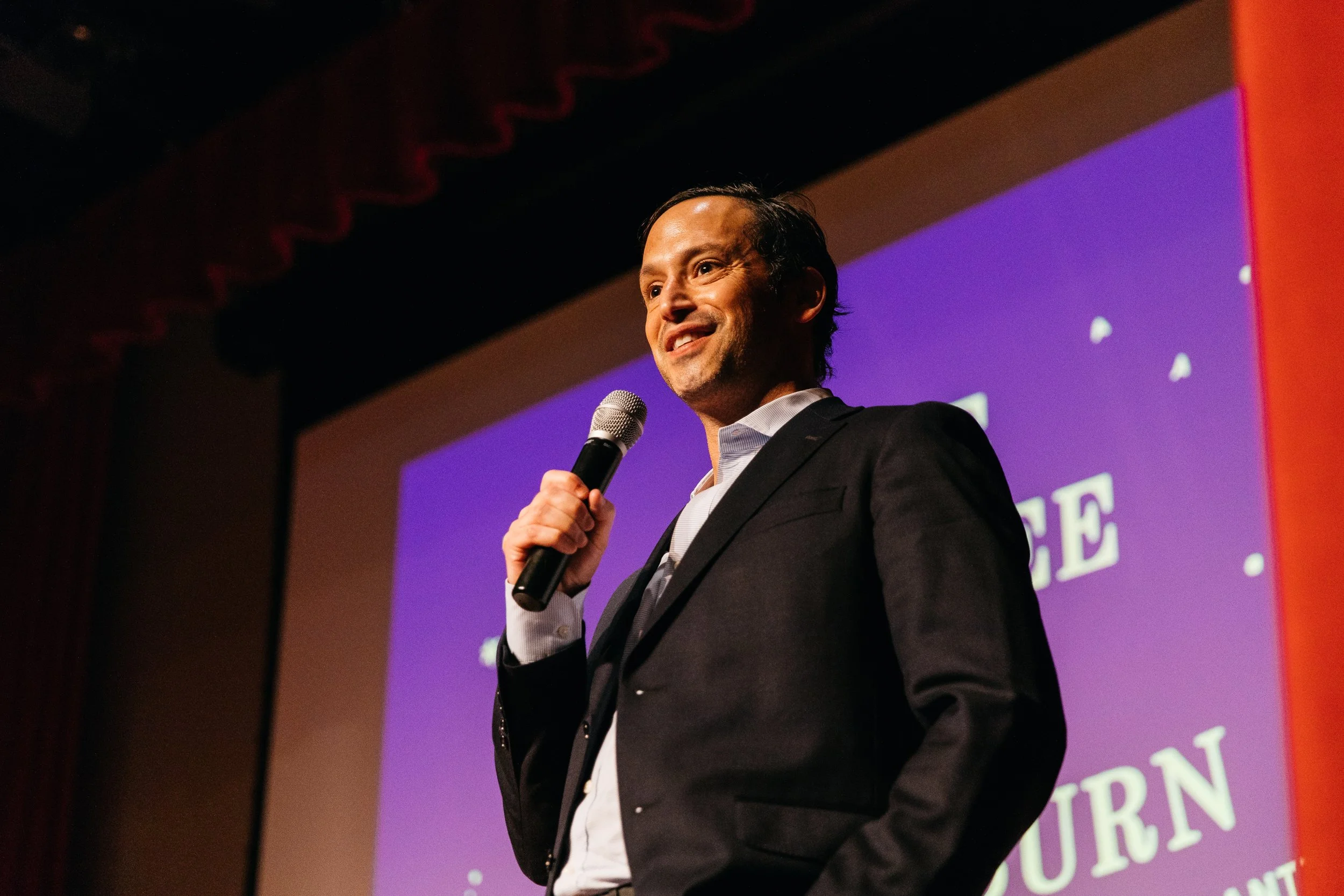A man in a suit holding a microphone and smiling during a presentation on stage with a purple background.