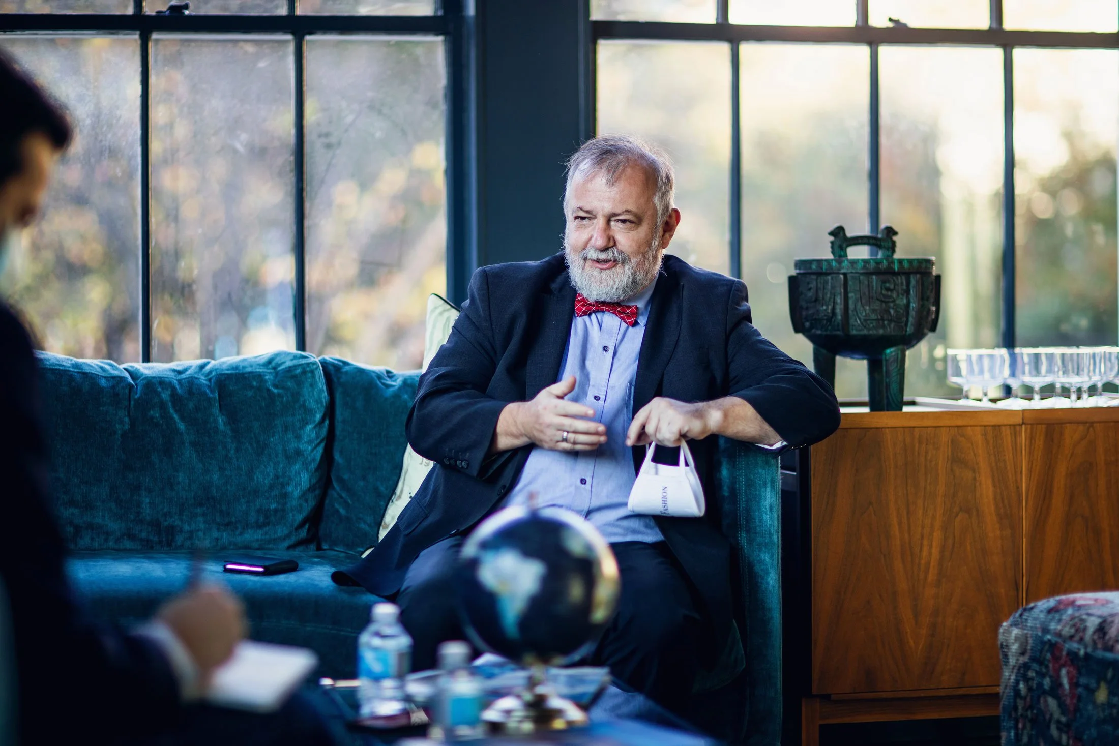 An elderly man with gray hair and a beard wearing a dark suit, light shirt, and red checkered bowtie, sitting on a blue couch in a brightly lit room with large window panes showing an outdoor scene with trees. He is engaged in conversation, gesturing