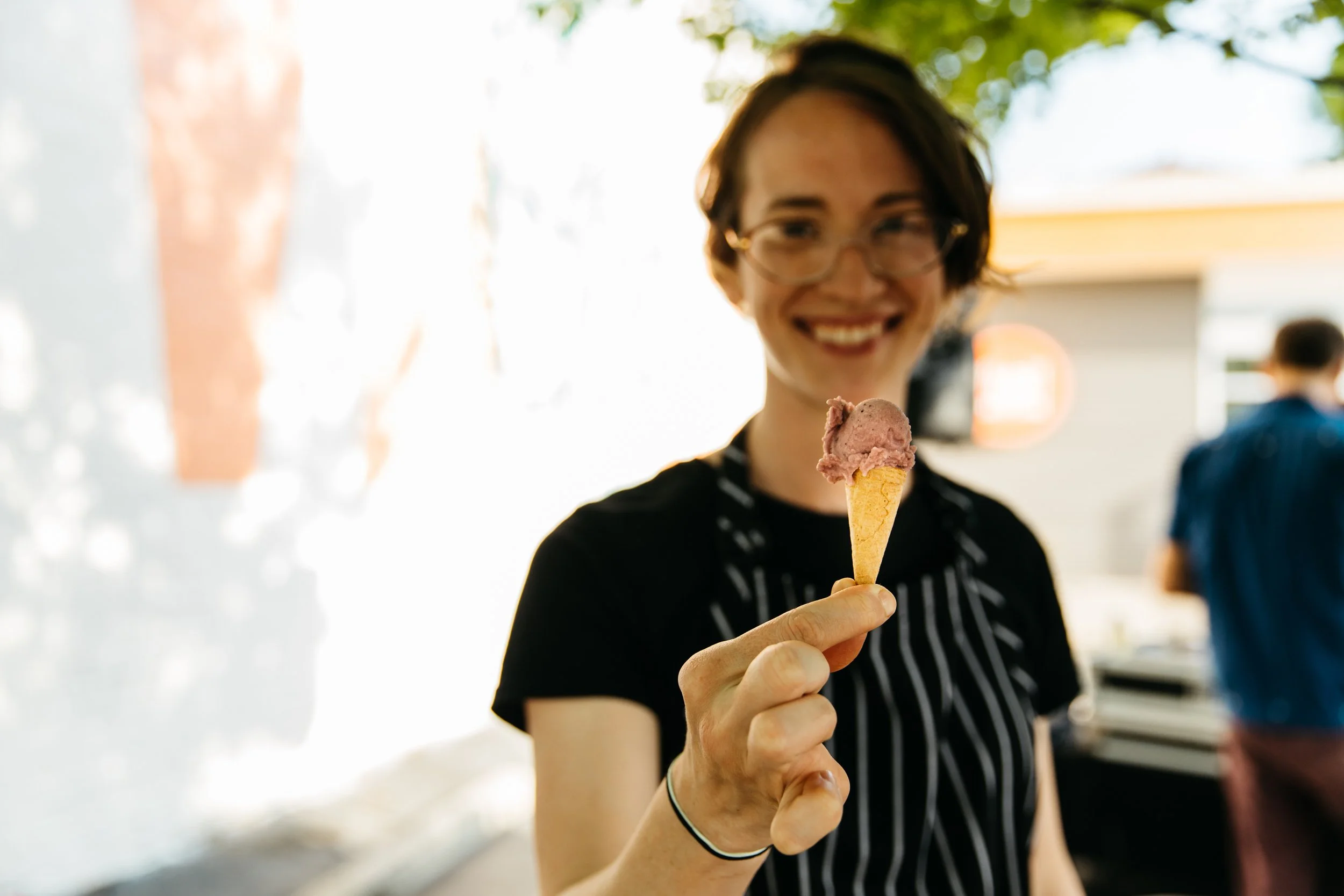 A young woman with short dark hair and glasses holding an ice cream cone with two scoops, smiling. She is wearing a black and white striped shirt and a black bracelet on her wrist.