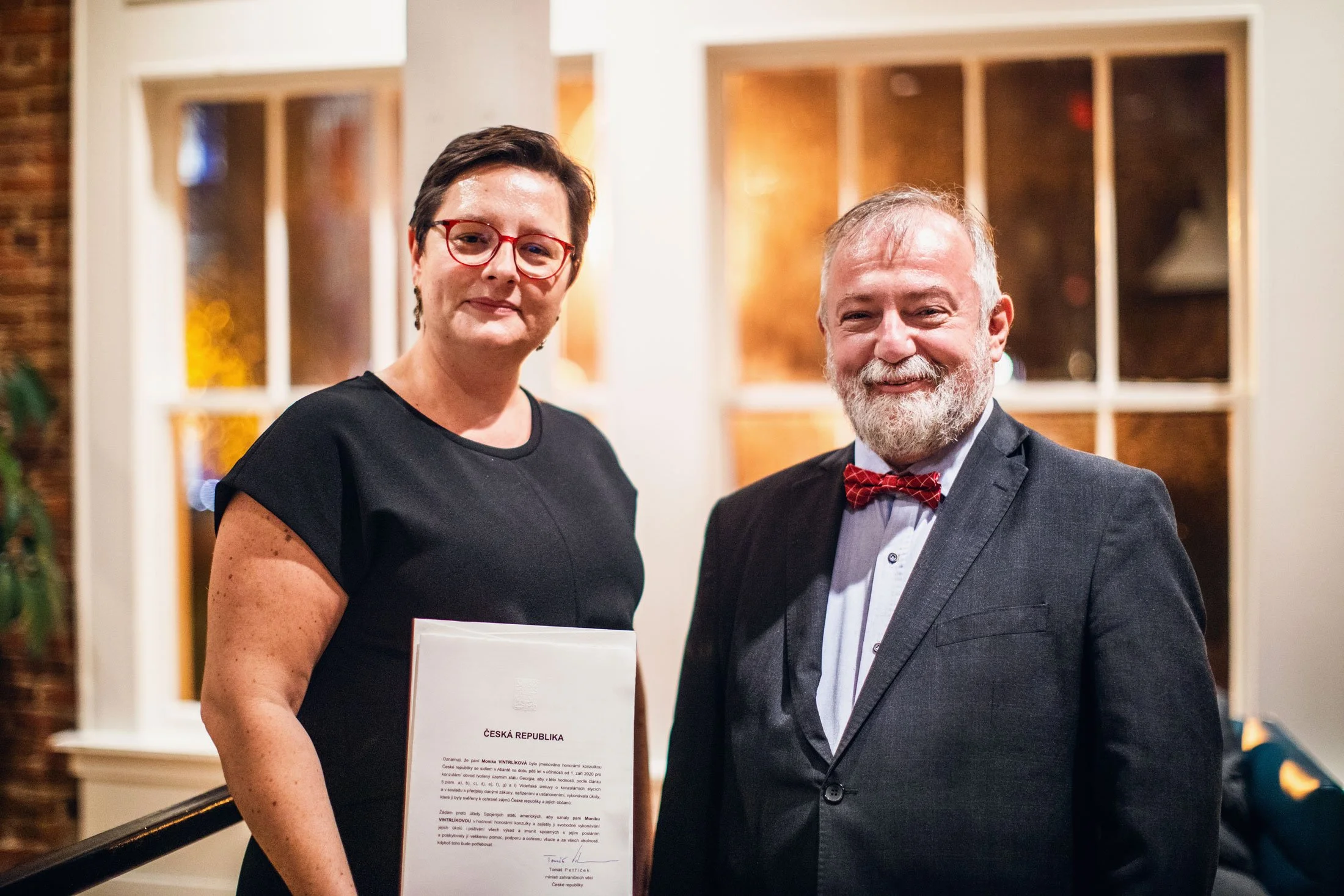 A woman with short dark hair, red glasses, and wearing a black dress is standing next to a man with gray hair, a gray beard, and dressed in a gray suit with a white shirt and red bow tie. They are both smiling and standing in front of a window with a wooden frame. The woman is holding a document with the words 'ČESKÁ REPUBLIKA' and additional text, and the background appears to be an indoor setting with warm lighting.