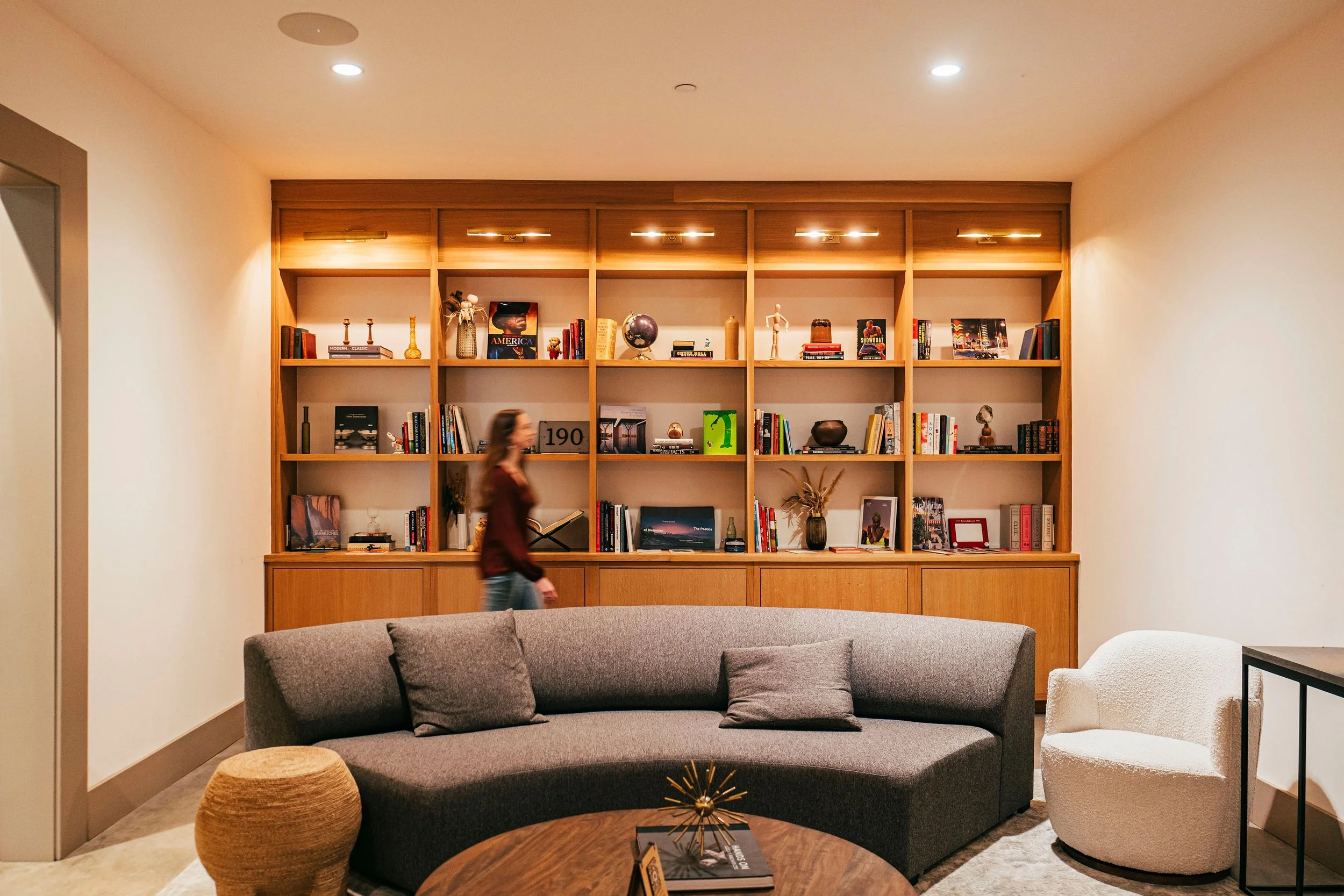 A contemporary living room features a matching dark gray sofa and a white textured armchair, with a wooden coffee table in the foreground. Behind the sofa, there is a large wooden bookshelf filled with books, decor items, and framed pictures. The roo
