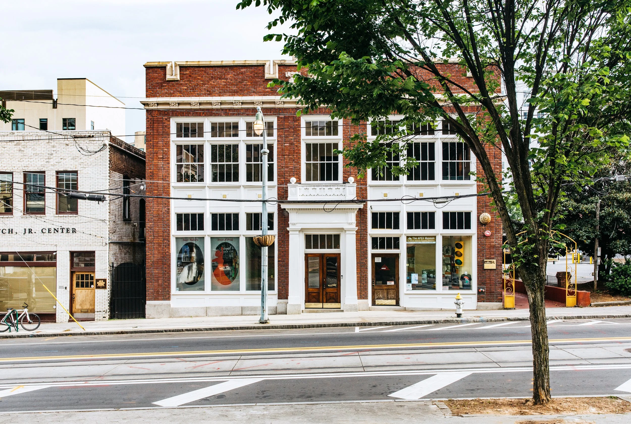 A historic brick building with large windows and a white decorative trim, located on a city street with a tree in the foreground and a streetlamp in front of the building.