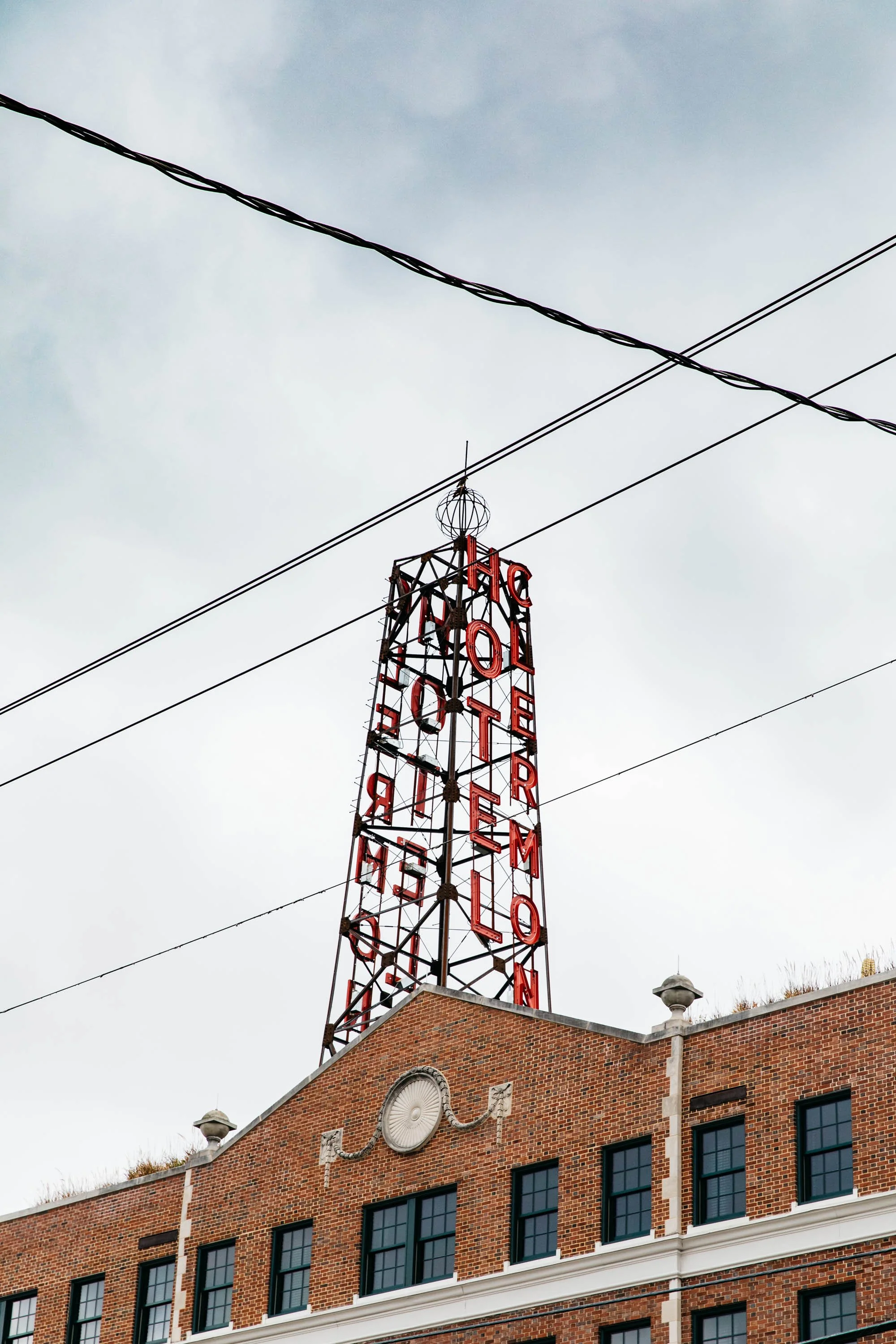 An old brick building with black-trimmed windows and decorative white architectural details, topped with a red neon sign saying "Hotel" and a metal tower with a sphere on top against an overcast sky.