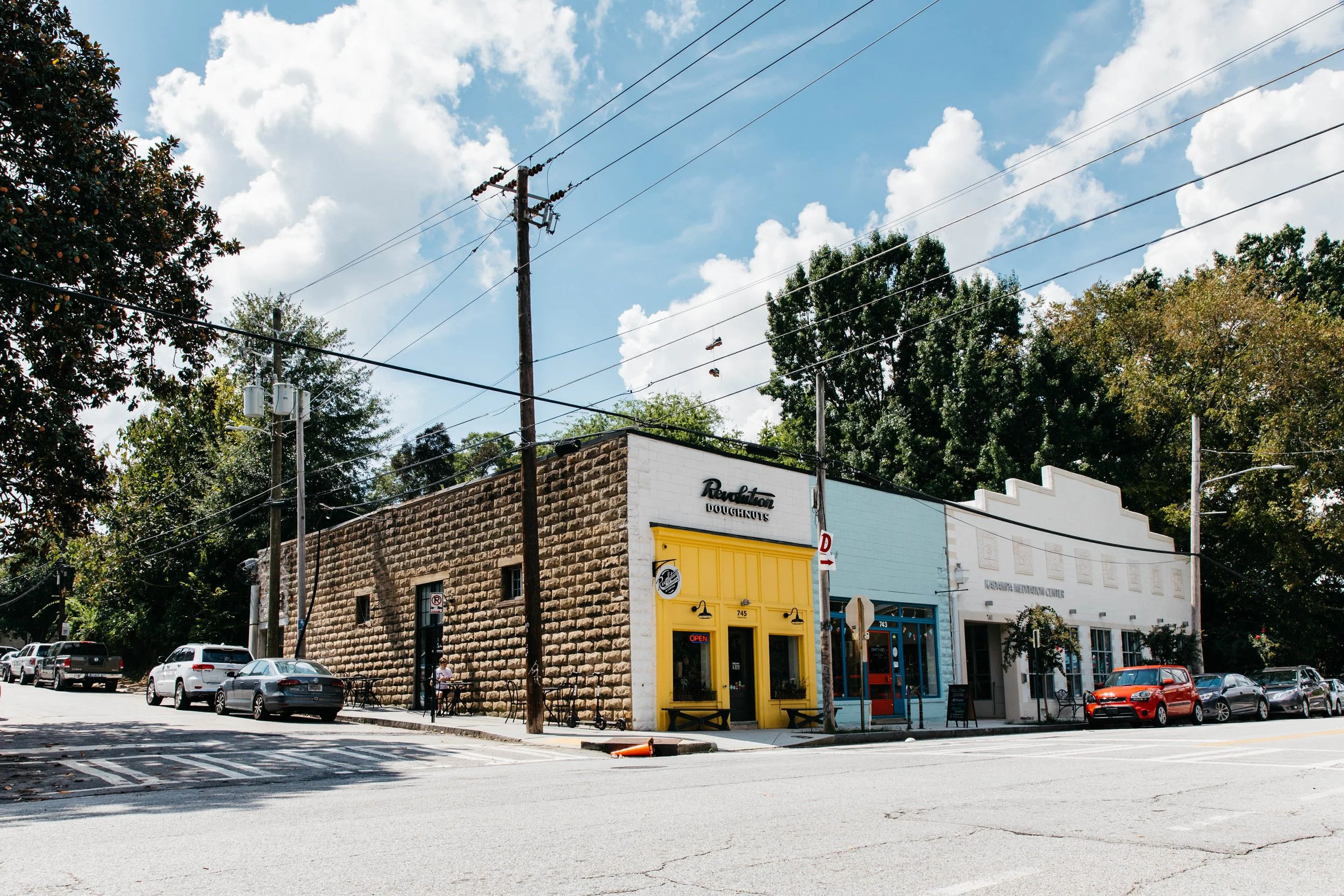 Street view of a small commercial building with yellow door and blue accents, parked cars on the street, utility poles with wires, trees, partly cloudy sky.