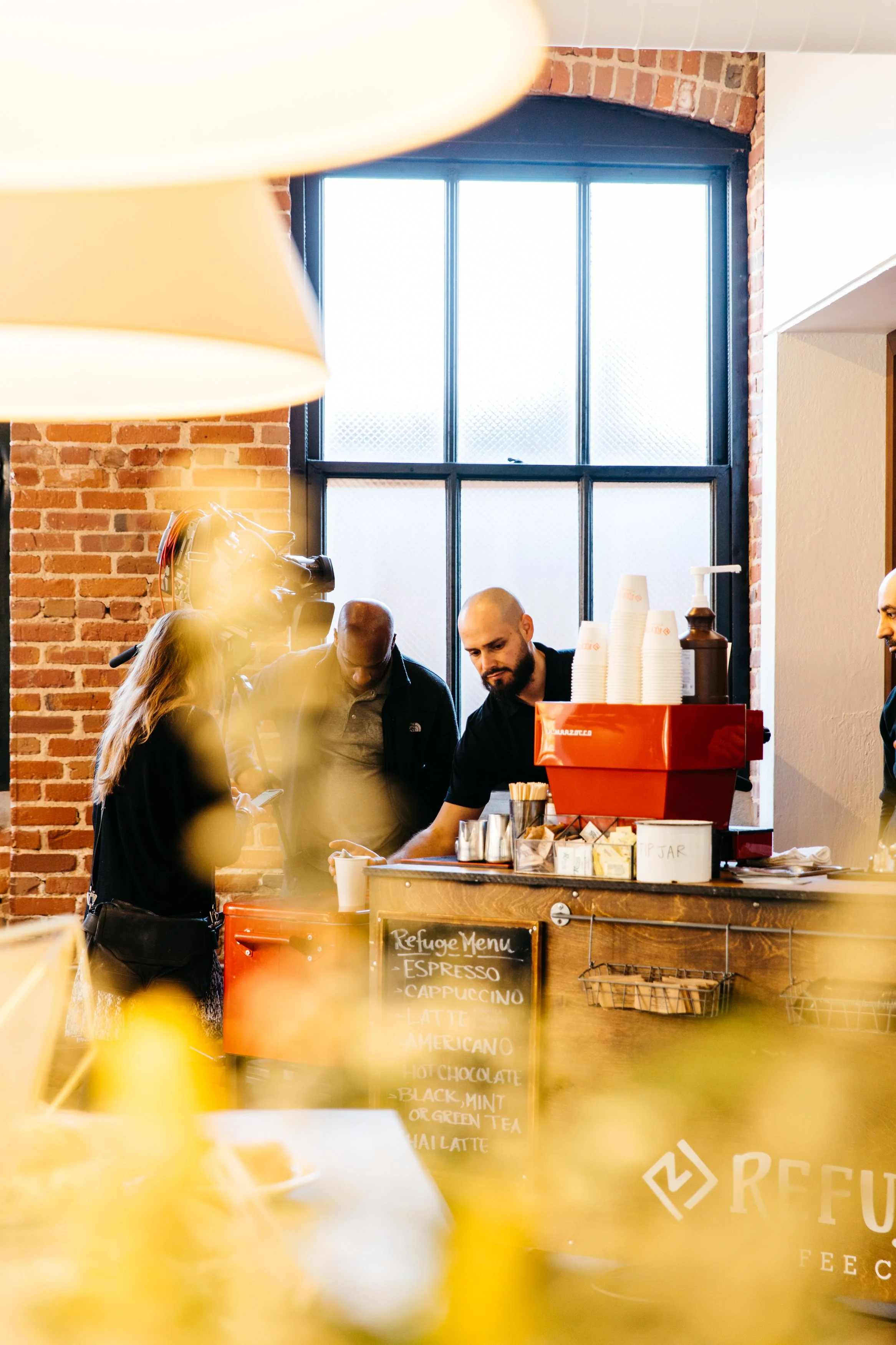 People ordering and being served coffee at a cafe counter with a brick wall and large window in the background.