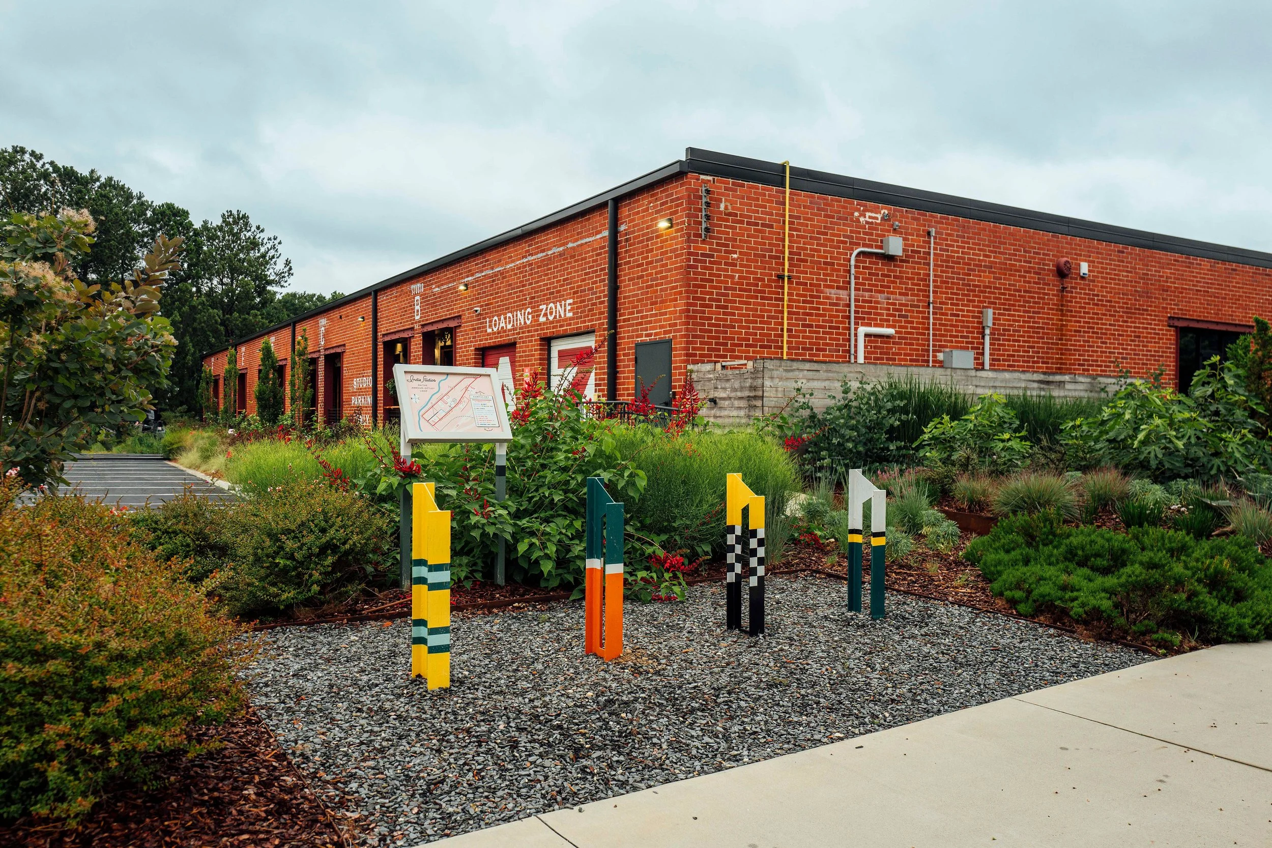 A red brick building with a black roof, surrounded by greenery and landscaped garden with yellow and black posts. The building has large garage doors and signs indicating loading zone and studio parking.