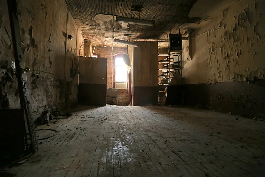 Empty, abandoned room with worn wood floor, peeling wall paint, an arched window letting in light, and various items like a radiator, a small shelf with speakers, and scattered debris.