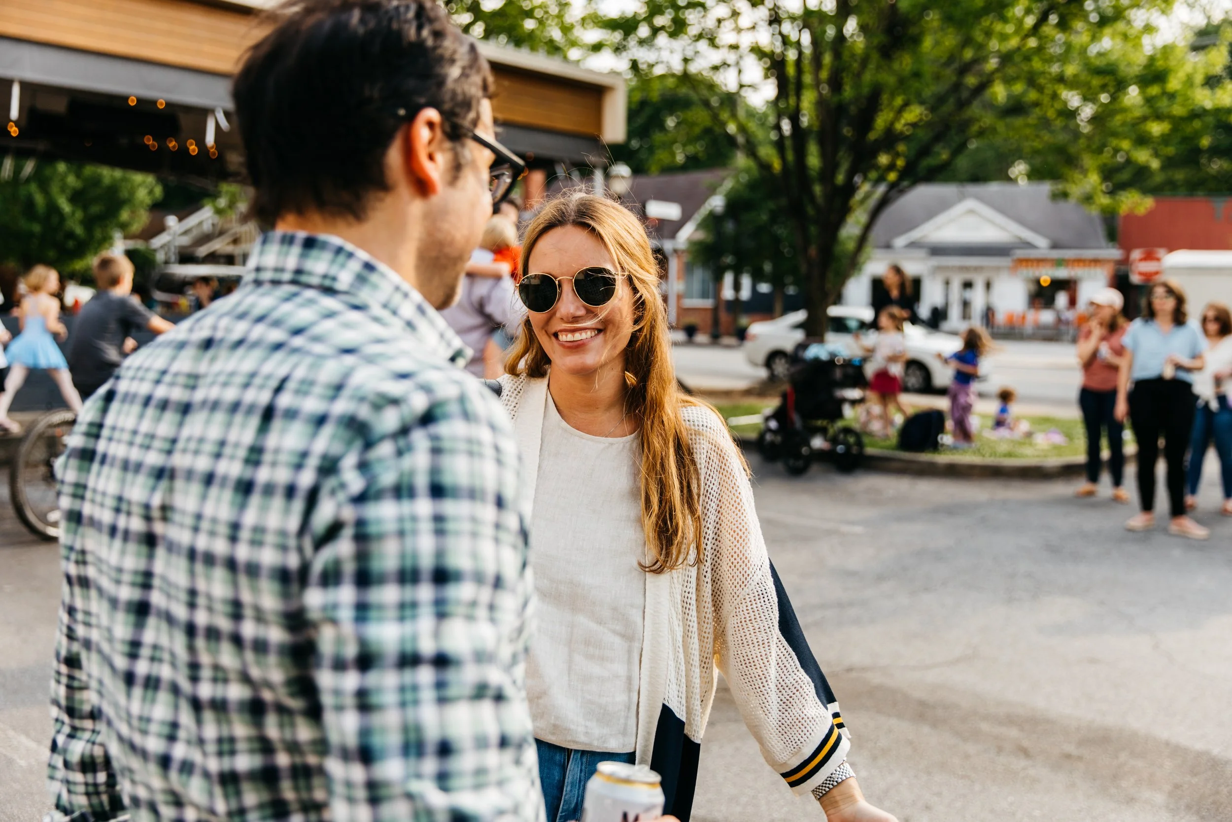 A woman with long red hair wearing glasses and a white sweater smiling and talking to a man with dark hair and glasses. They are outdoors in a lively area with other people in the background, some walking and others talking, with trees and buildings 