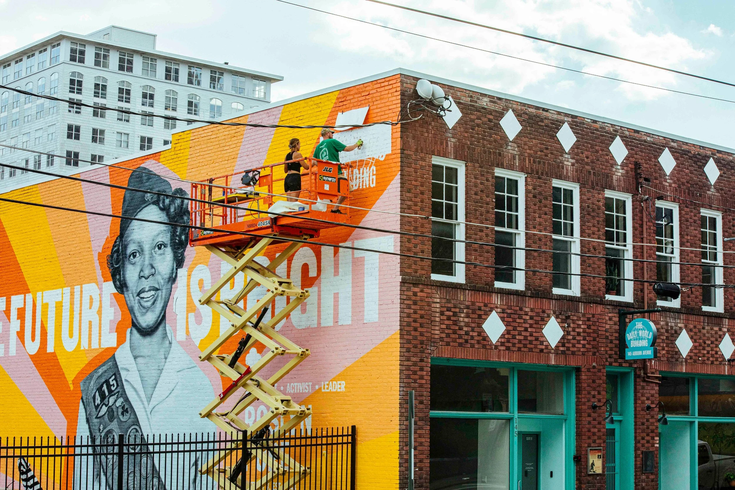 Two people on an orange scissor lift painting a mural on a brick building wall. The mural features a woman with short curly hair, wearing a shirt with glasses, and holding a book or folder.