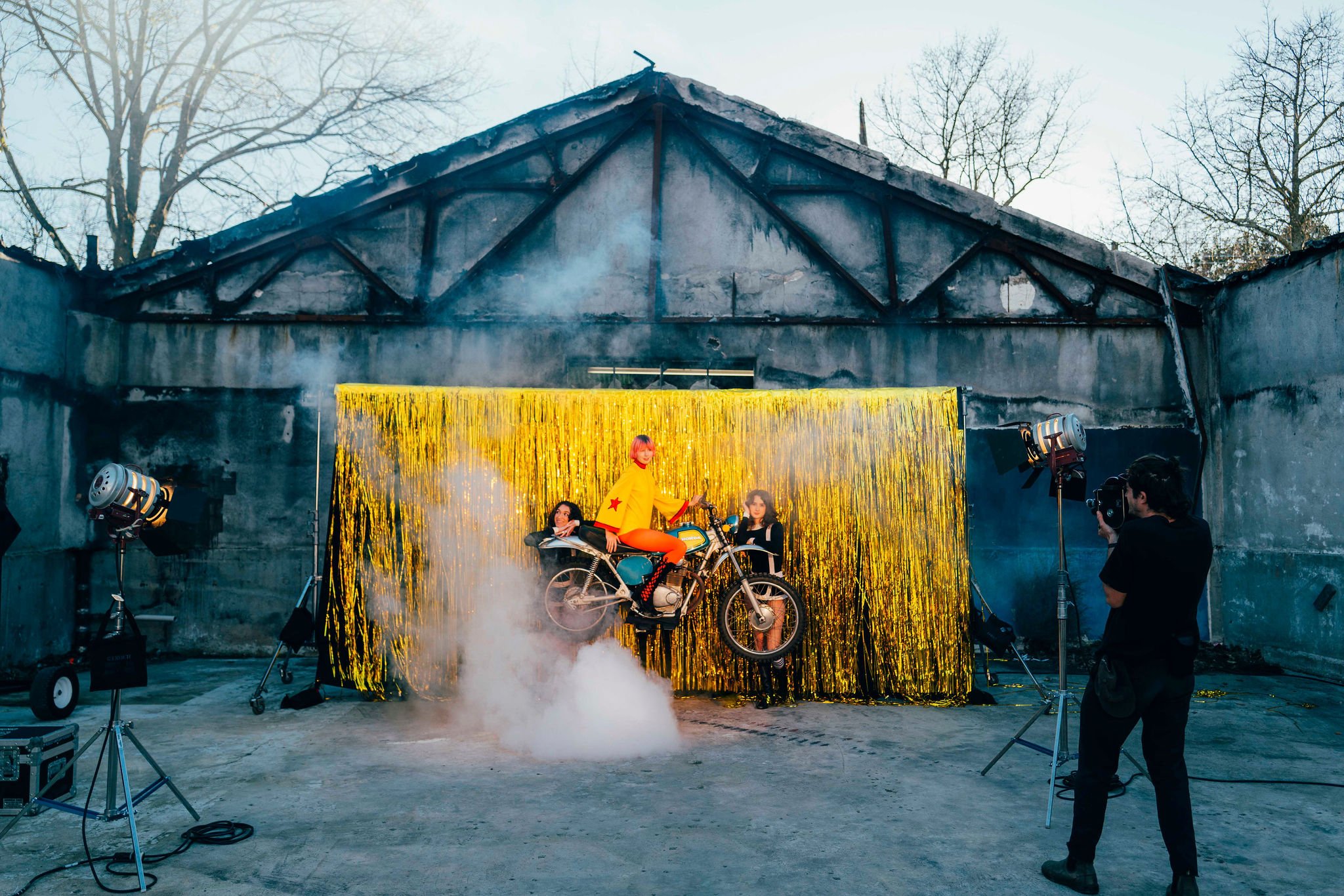A group of models posing on a motorcycle during a photo shoot outdoors in front of a yellow shimmering backdrop, with professional lighting and camera crew capturing the scene.