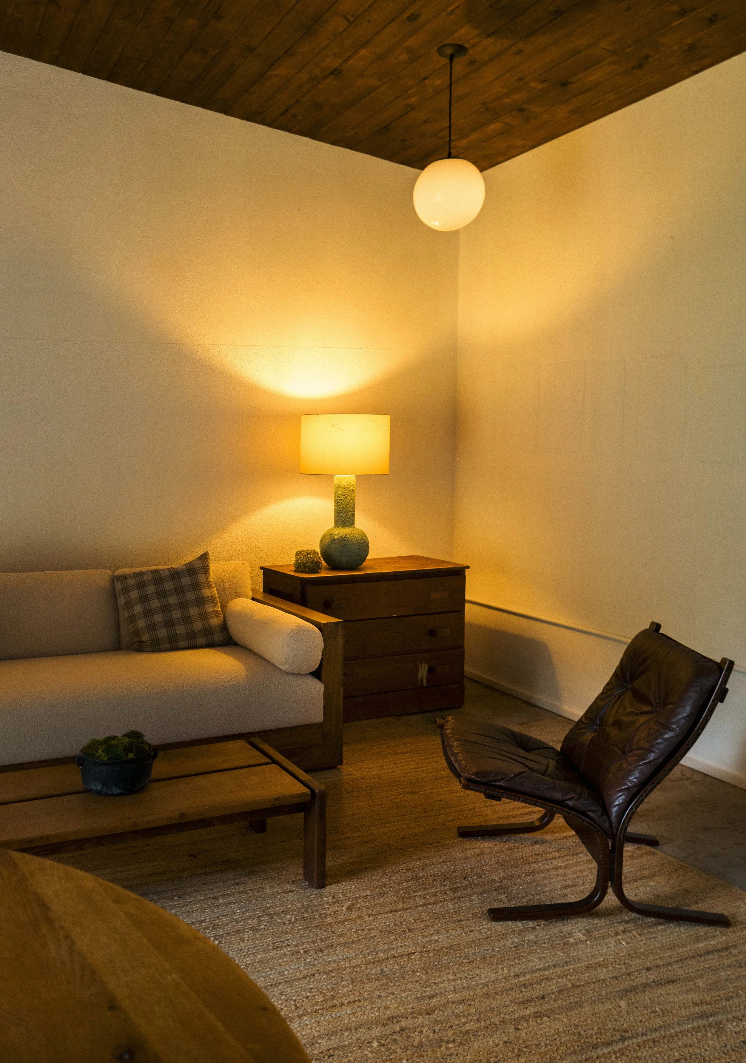 Living room with beige sofa, wooden coffee table, table lamp, and black leather chair, with a wood-paneled ceiling and warm lighting.