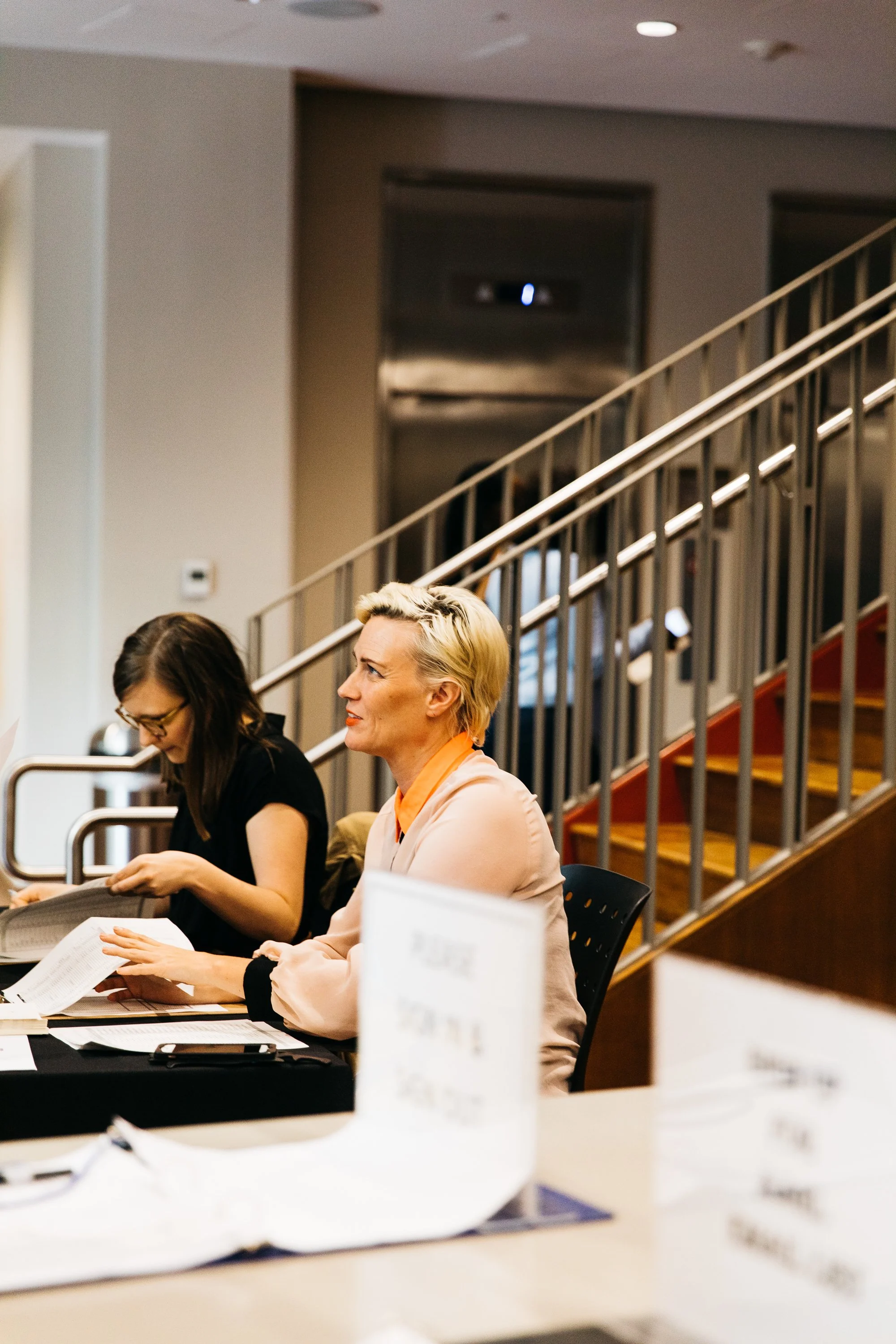 Two women sitting at a conference table with papers and pens, engaged in conversation, in a room with a gray staircase and railing in the background.