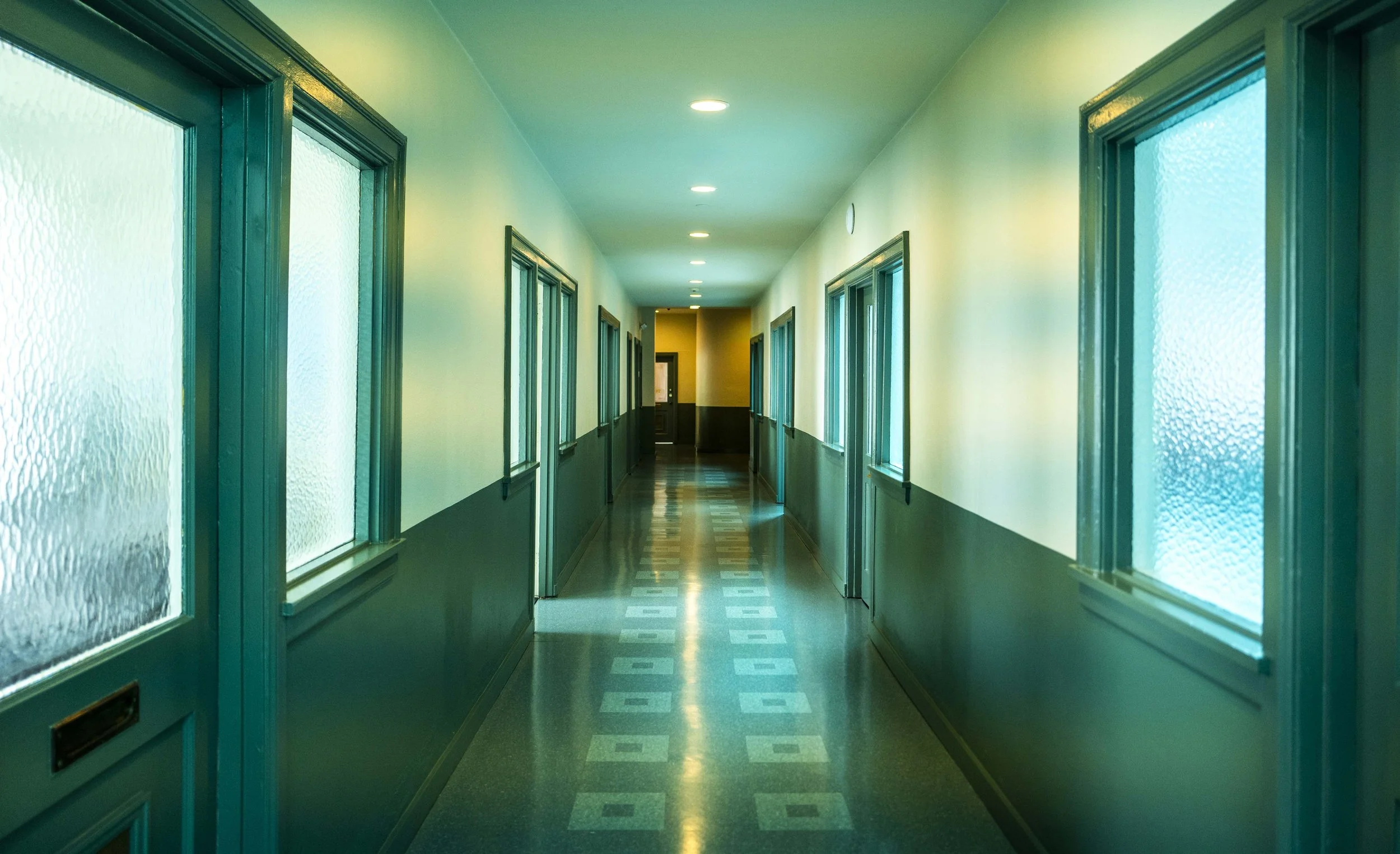 Long hallway with windows on both sides, green walls, wood-paneled lower half, and patterned tile floor, illuminated by ceiling lights.