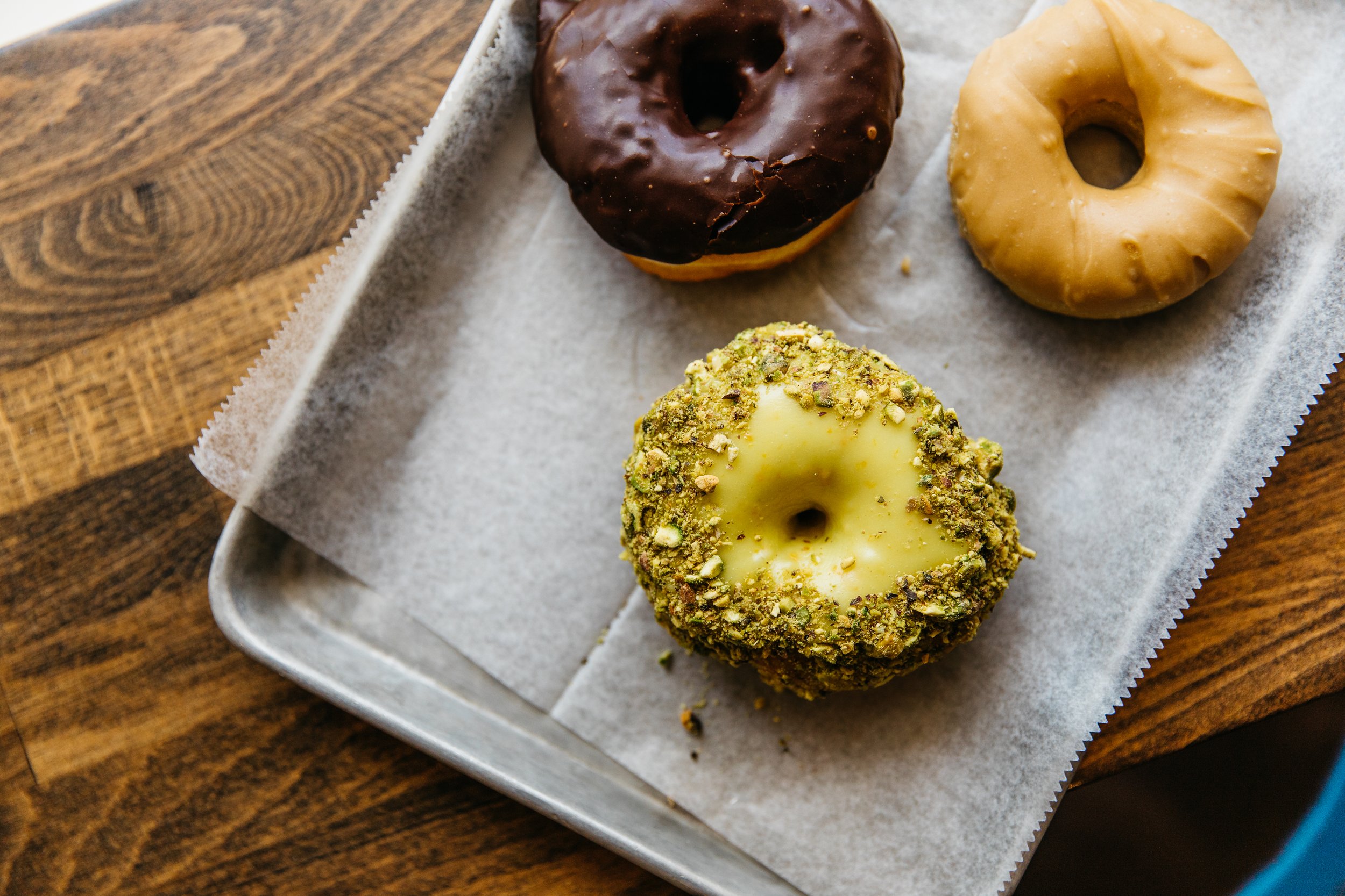 Three donuts on a parchment paper-lined tray, with a wooden surface beneath. One donut is chocolate glazed, another is caramel glazed, and the third is coated with crushed pistachios.
