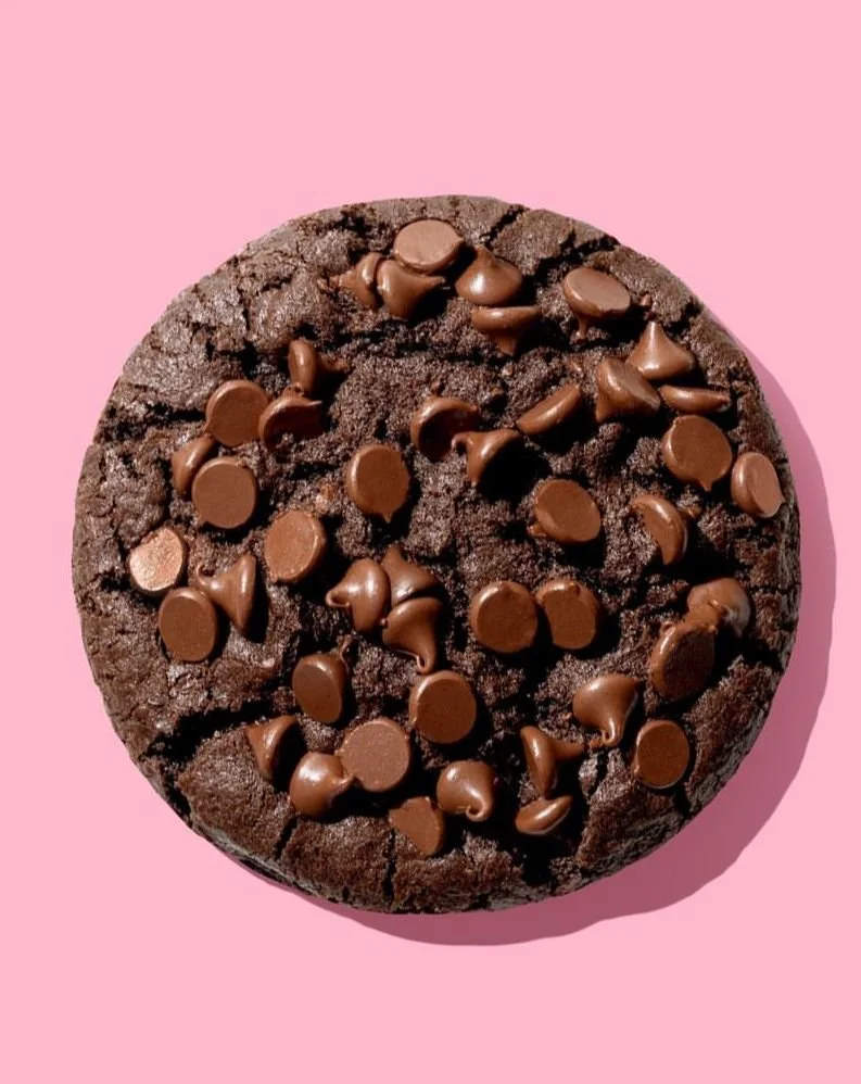 A round chocolate cookie topped with melted chocolate chips on a pink background.